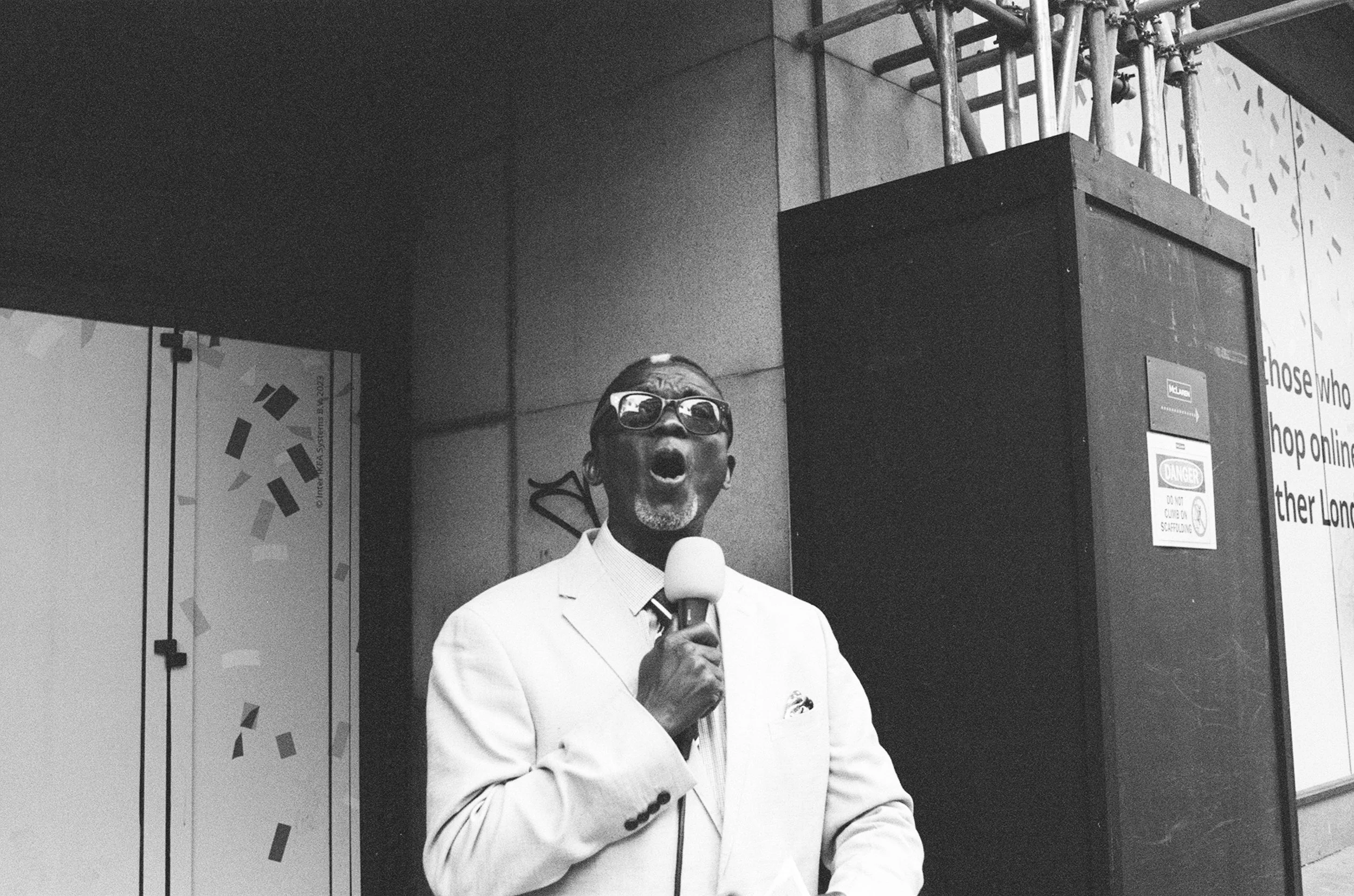 A man in a white suit and sunglasses singing or speaking into a microphone in an indoor setting.