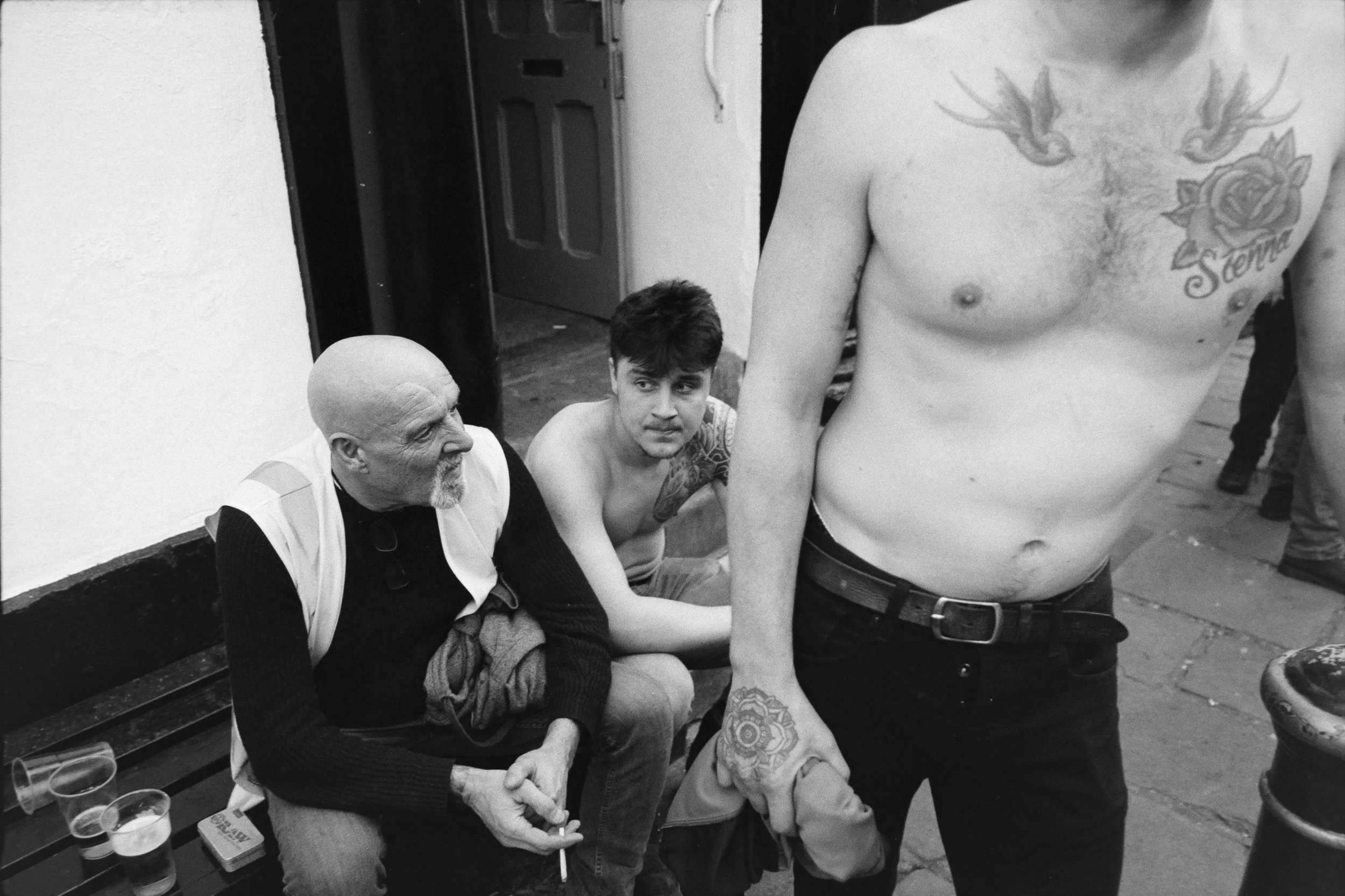 A black and white photo showing two shirtless men with tattoos sitting on a bench, and an older man sitting on a bench nearby. The older man has a shaved head and a goatee, while one of the shirtless men has dark hair and a tattoo on his chest.