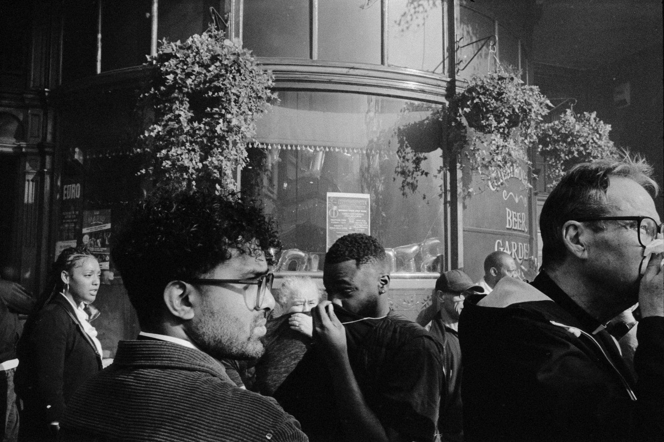Group of people standing outside a pub with hanging flower baskets, some are holding tissues or breathing masks, in black and white photo.