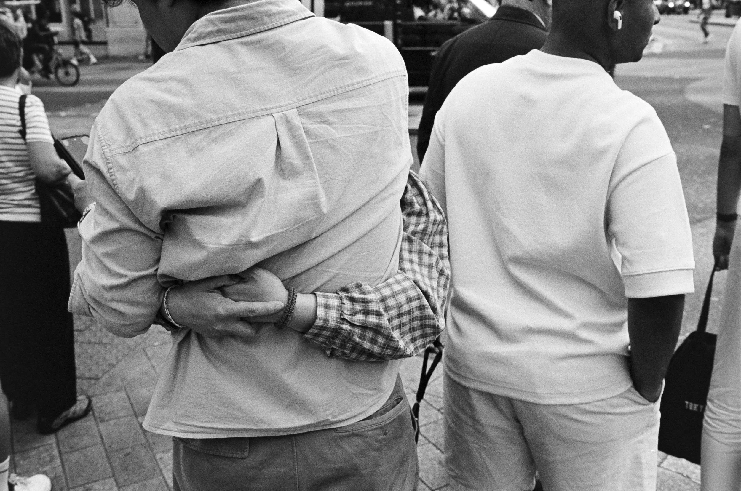 People standing on a city sidewalk, some with hands in pockets or arms crossed, wearing casual clothing.