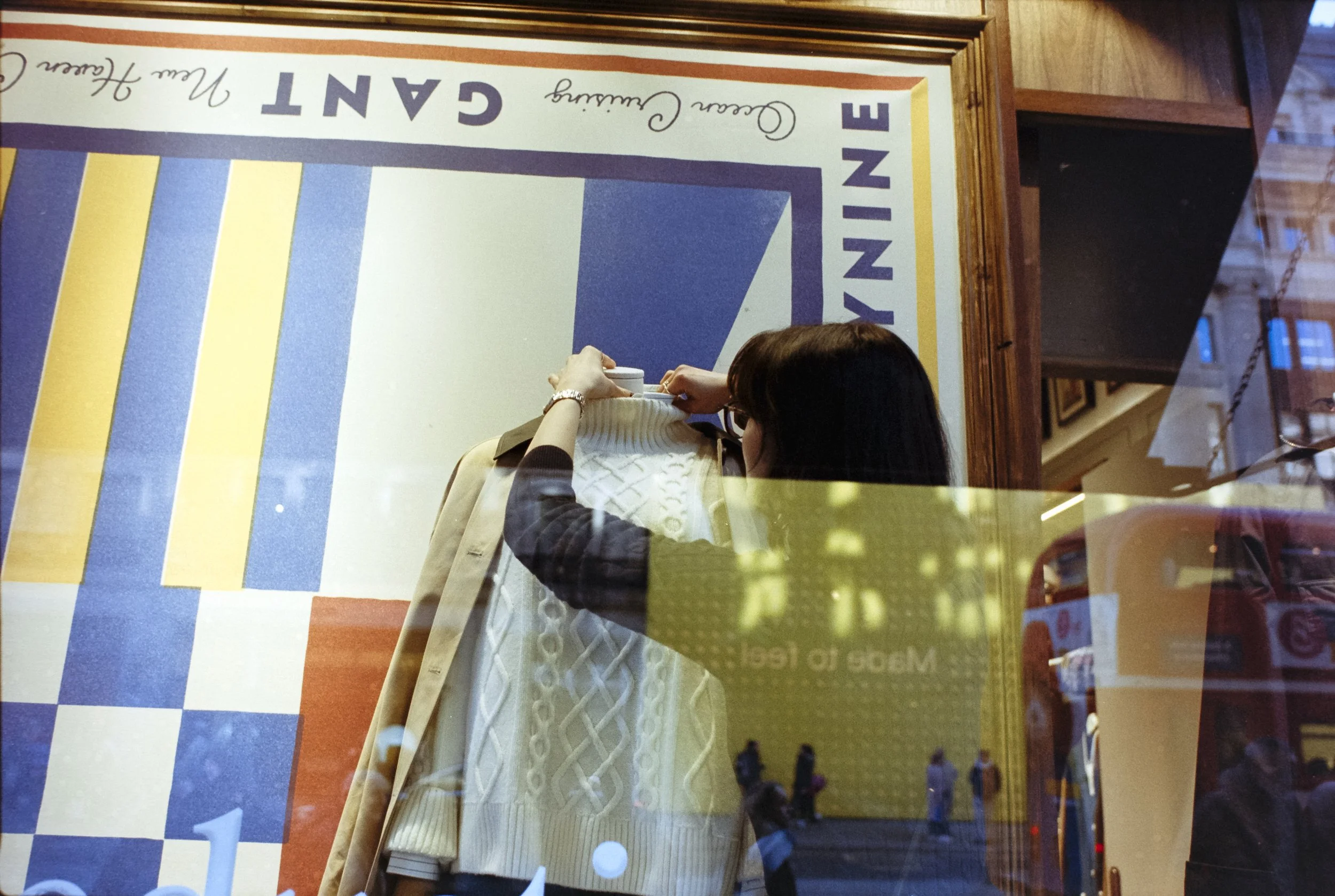 A woman with dark hair and glasses hanging inside a restaurant or cafe, arranging or inspect a sweater on a mannequin, seen through a window with reflections of pedestrians and street scene outside.