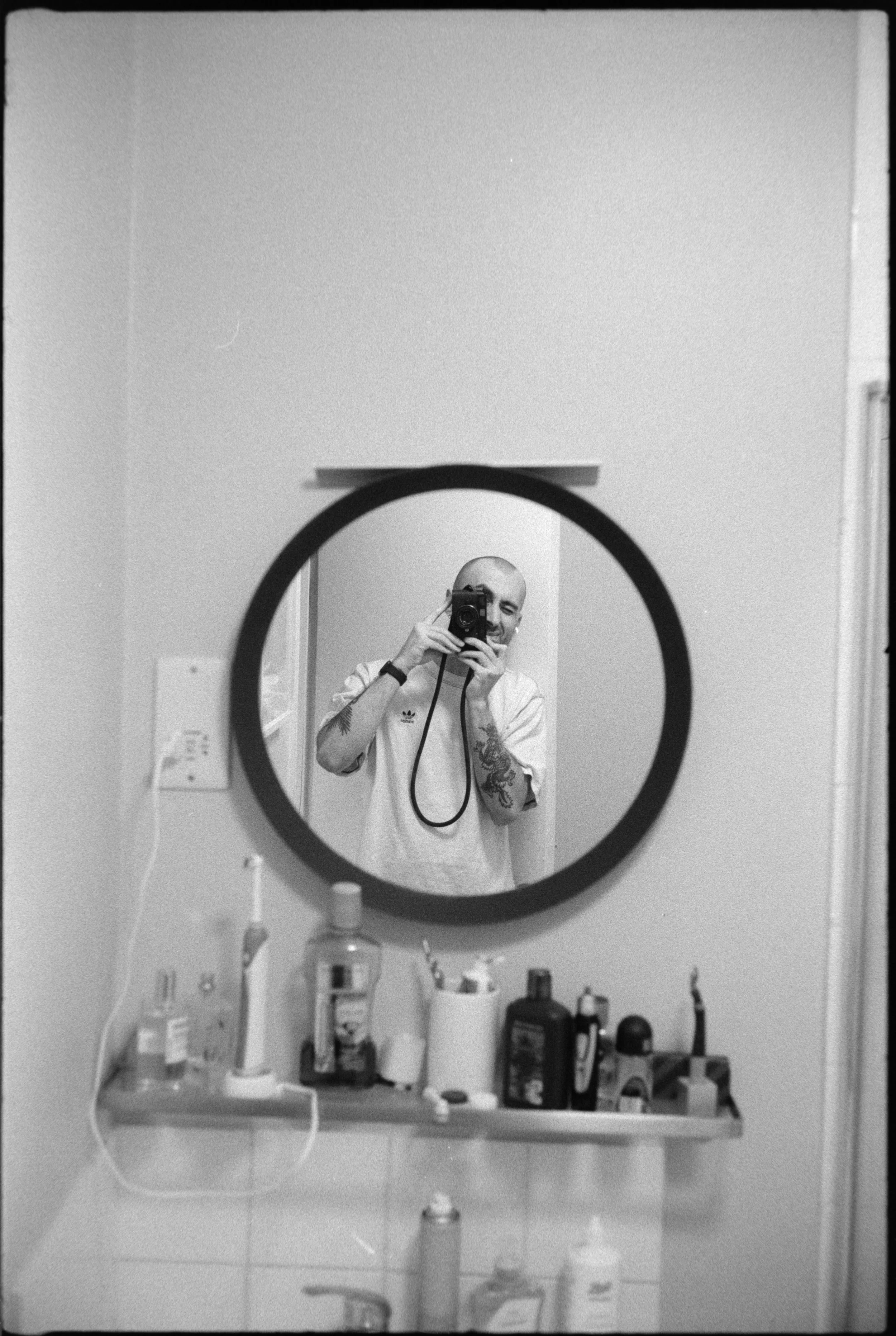 A black and white photo of a person taking a mirror selfie in a bathroom. The person has a shaved head, is smiling, and is wearing a light-colored shirt with a logo on the left side. There are various bathroom products on the shelf beneath the mirror.