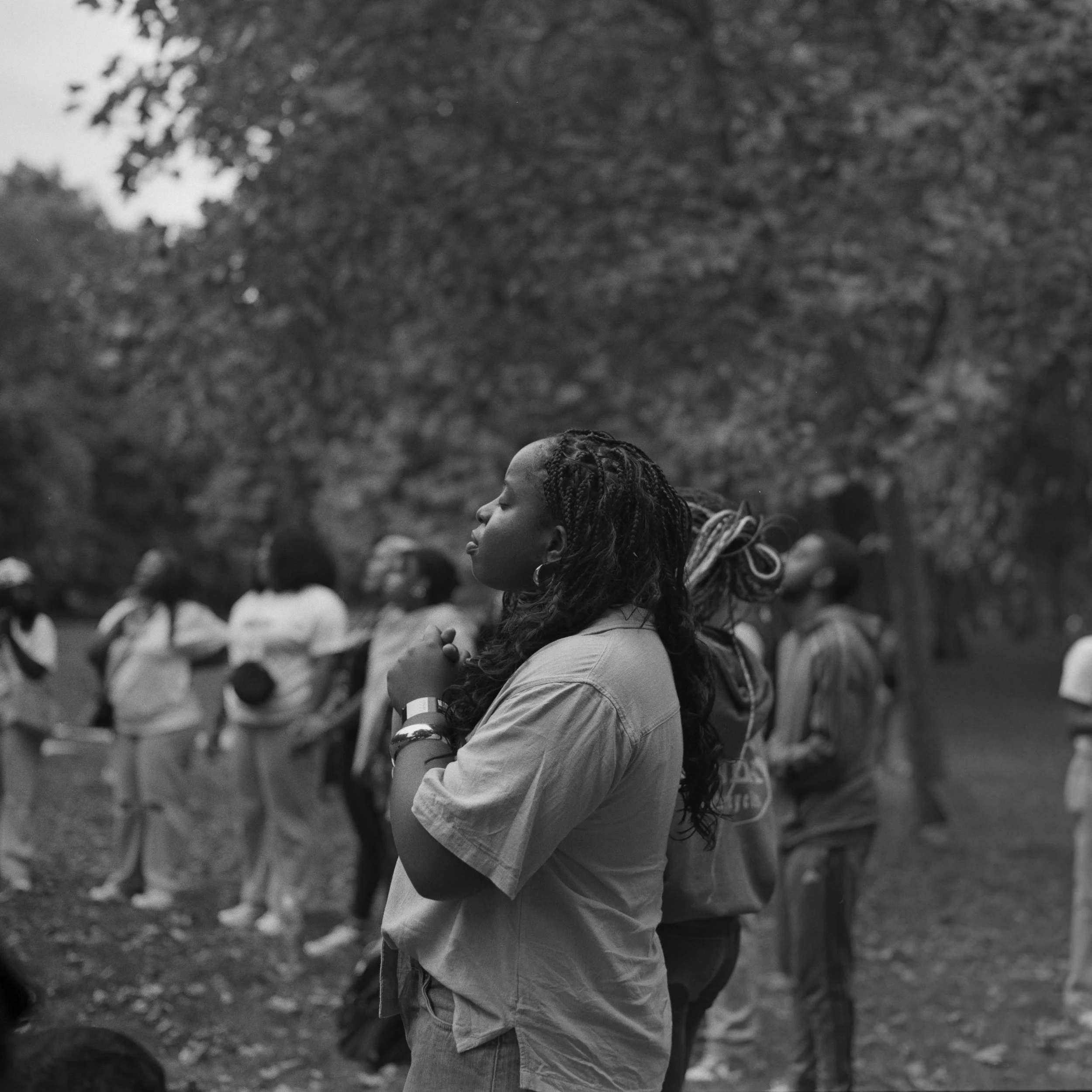 A black-and-white photo of a young woman with braided hair, standing with her eyes closed and hands clasped in front of her, in an outdoor gathering with other people and trees in the background.