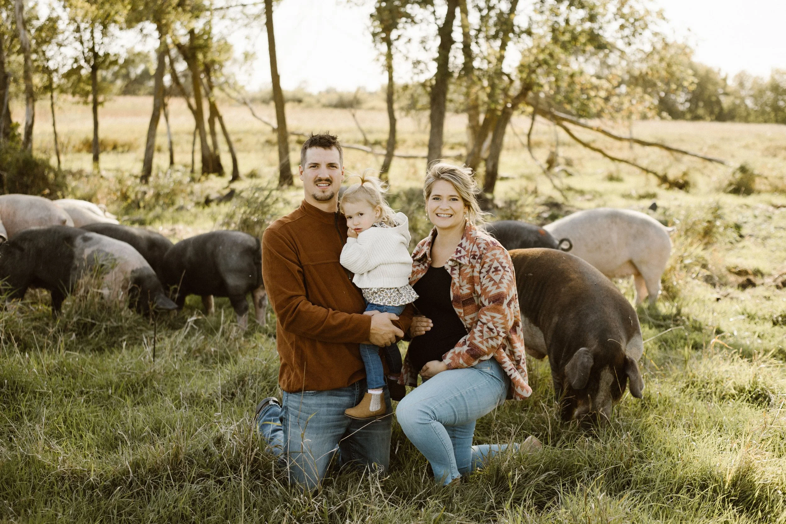 Kneeling family with two adults and a child, outdoors in a field with grazing pigs and trees in the background, during daytime.