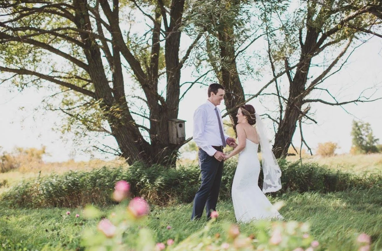 A couple in wedding attire holding hands outdoors, standing on green grass with a large tree and blue sky in the background.