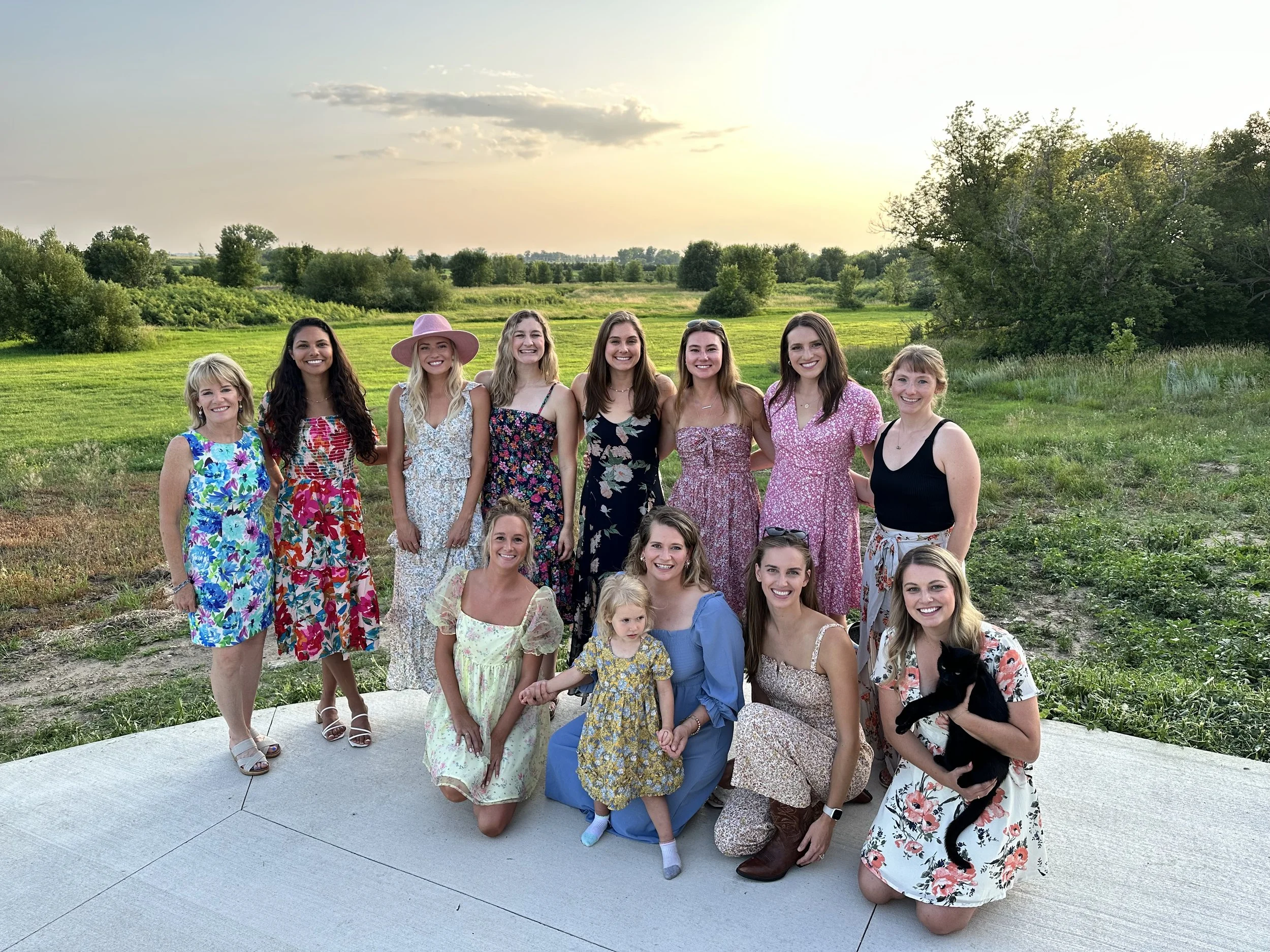 Group of women and a young girl outdoors during sunset, standing on a paved area with grass and trees in the background. They are smiling and dressed in summer dresses.