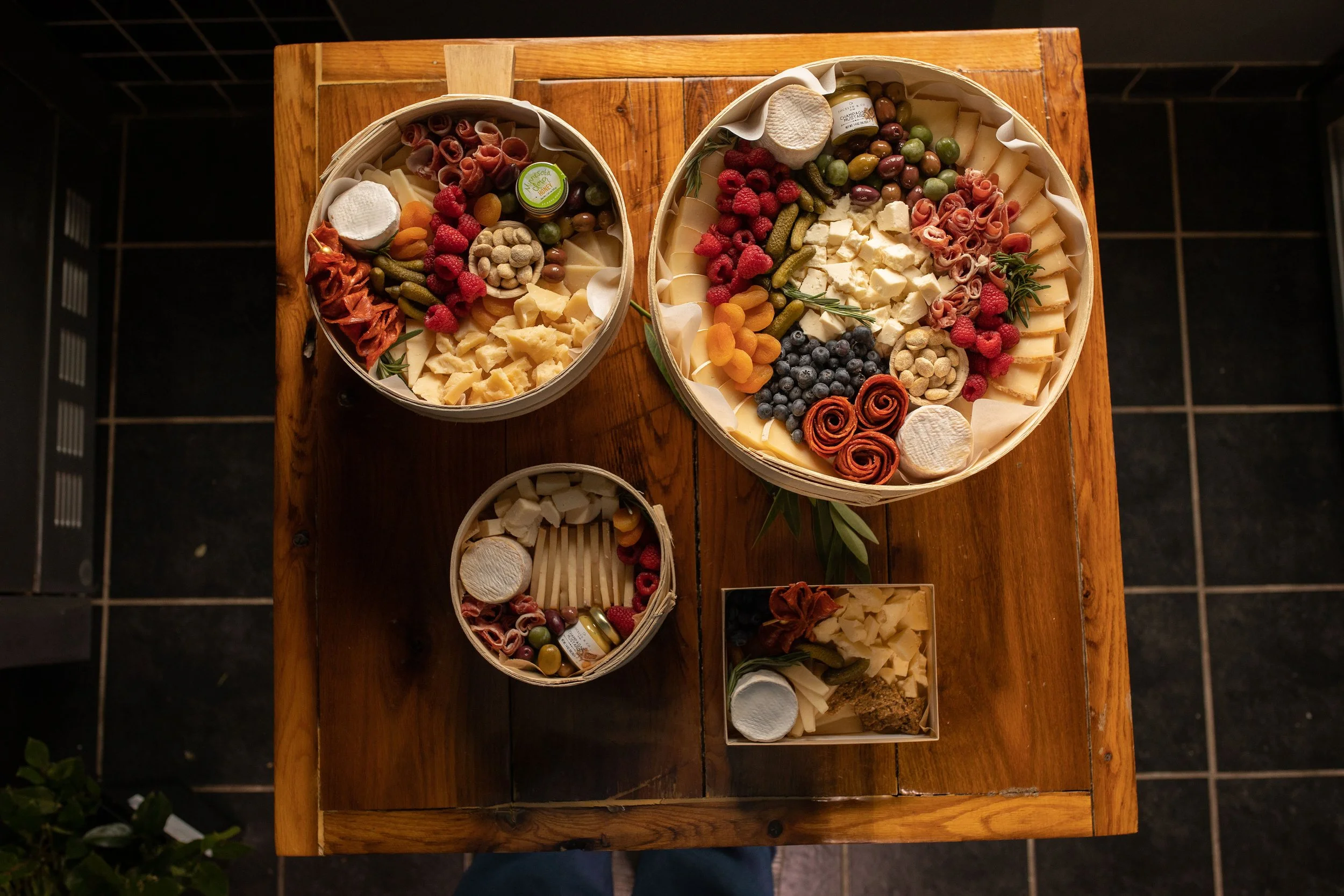 Assorted cheeses, cured meats, breadsticks, grapes, raspberries, blueberries, olives, and small jars of condiments arranged on a wooden table in round and rectangular baskets.