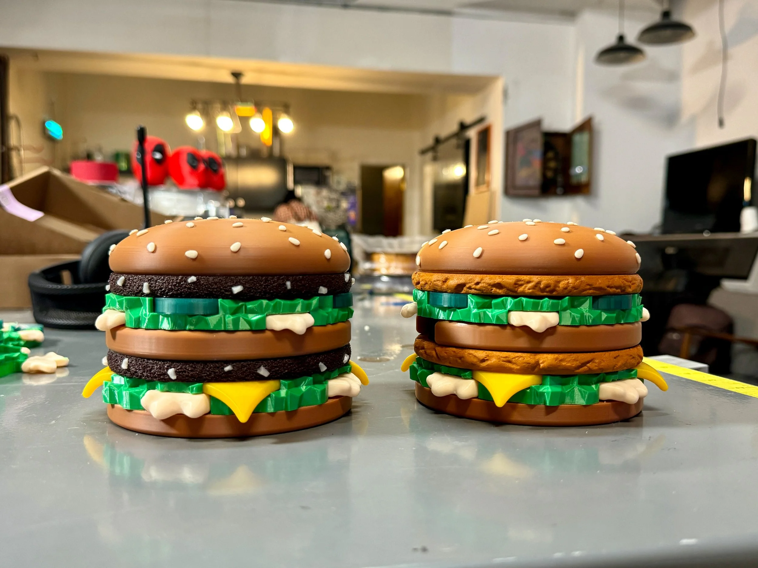 Close-up of two toy hamburger stacks with sesame seed buns, lettuce, cheese, onions, pickles, and meat, placed on a table in a kitchen or dining area.