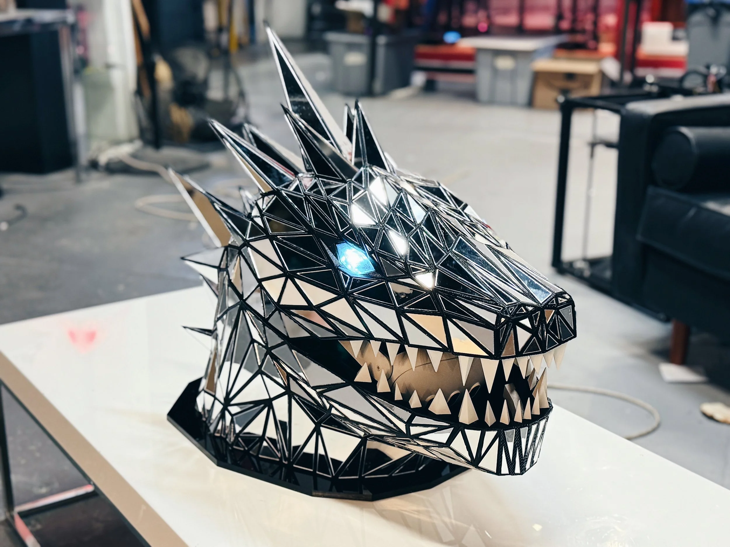 A geometric sculpture of a wolf's head made of reflective black and silver materials, placed on a work surface in a workshop or studio environment.