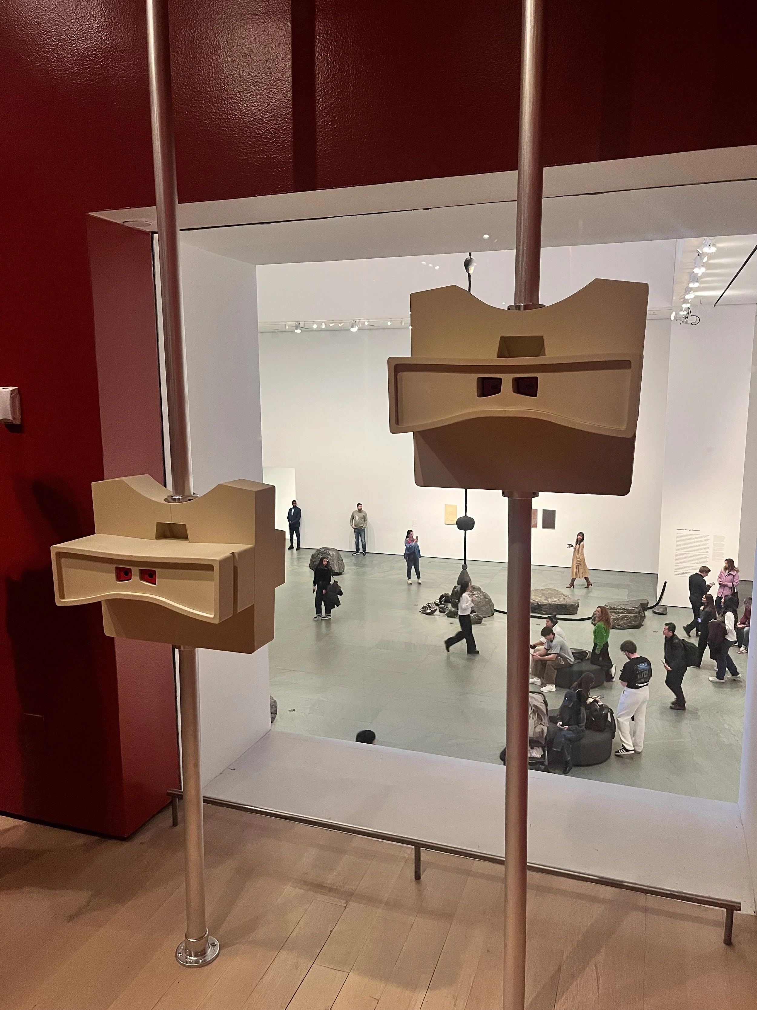 View from a bathroom window into an art gallery showing visitors walking and sitting in a large, open space with rocks on the floor, white walls, and spotlights on the ceiling.