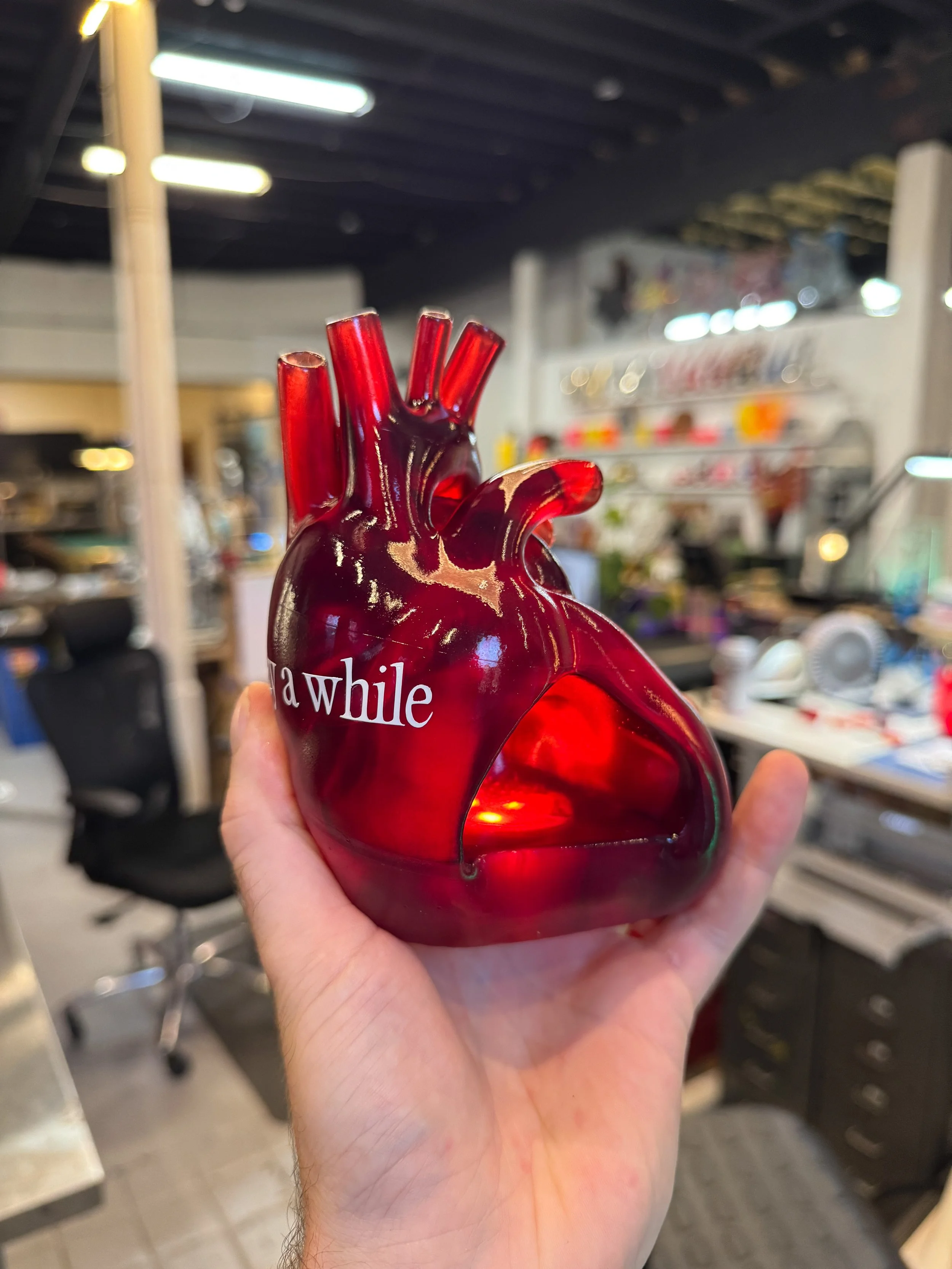 A person holding a red, anatomically-shaped glass heart sculpture with the word "awhile" written on it, in a room with shelves and blurred objects in the background.