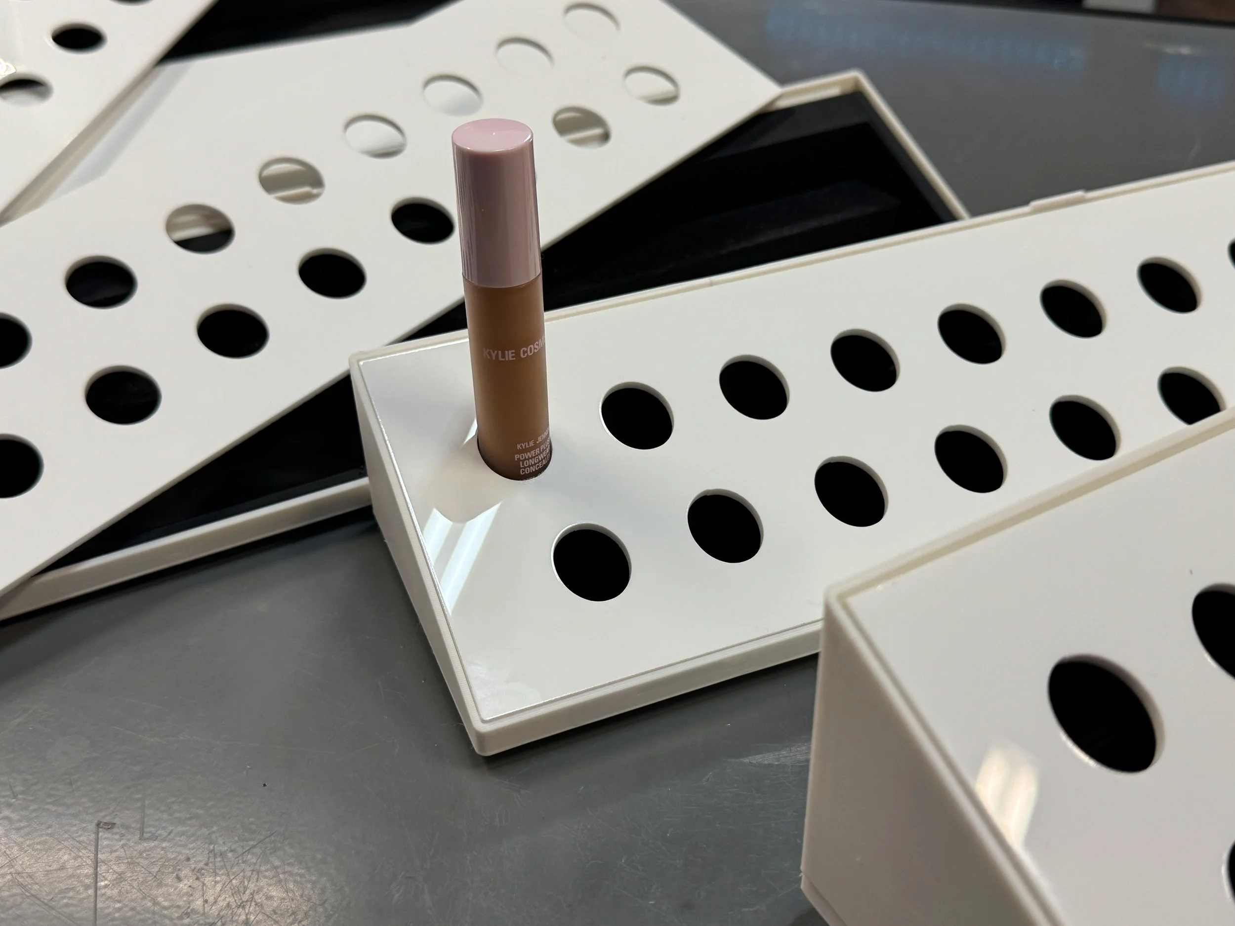 A Kylie Cosmetics lip product standing upright in a white display stand with black dot openings, outdoors on a gray surface.