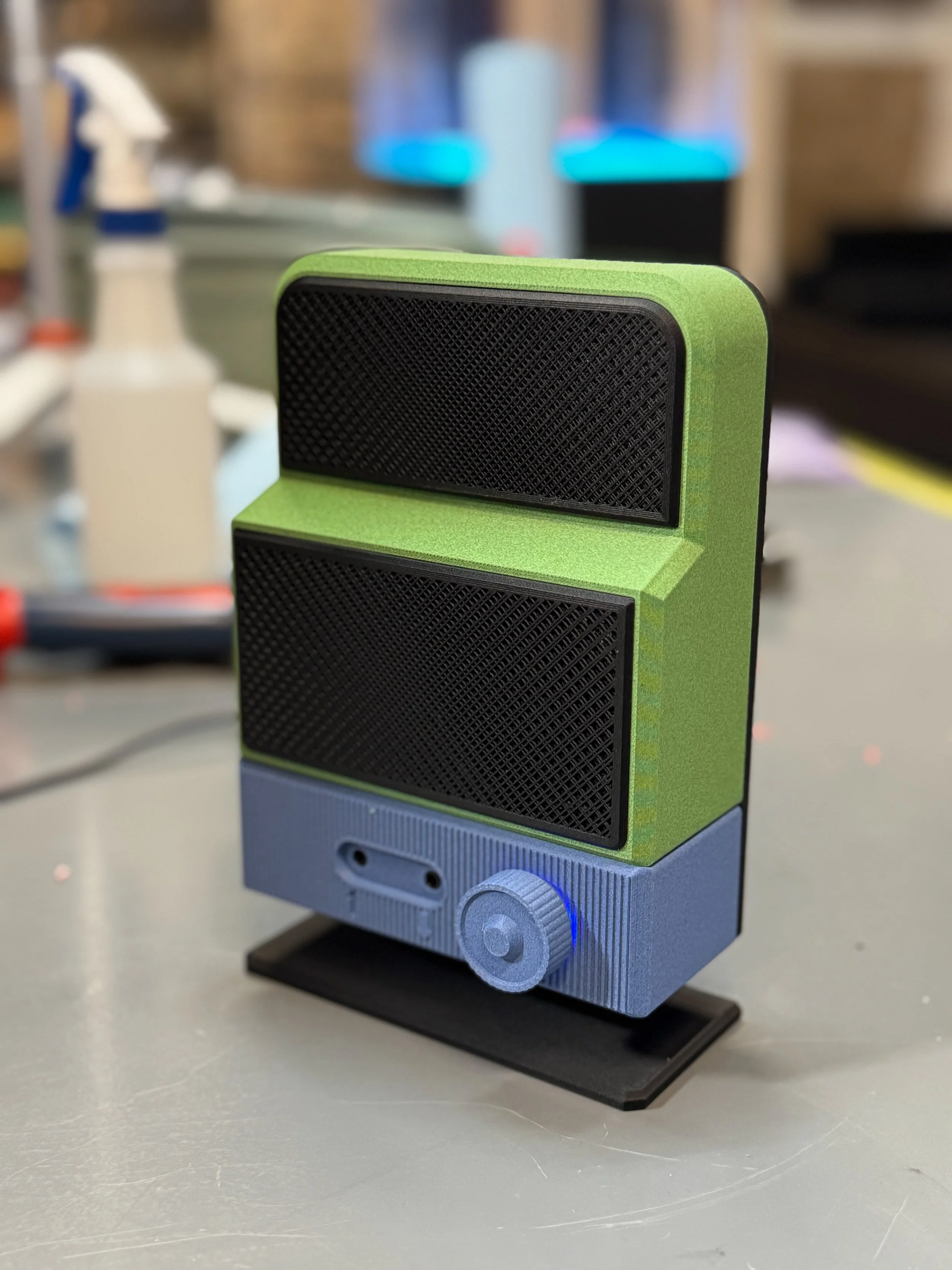 Close-up of a small portable speaker with a green and gray exterior and black speaker grilles, placed on a stand on a gray surface.