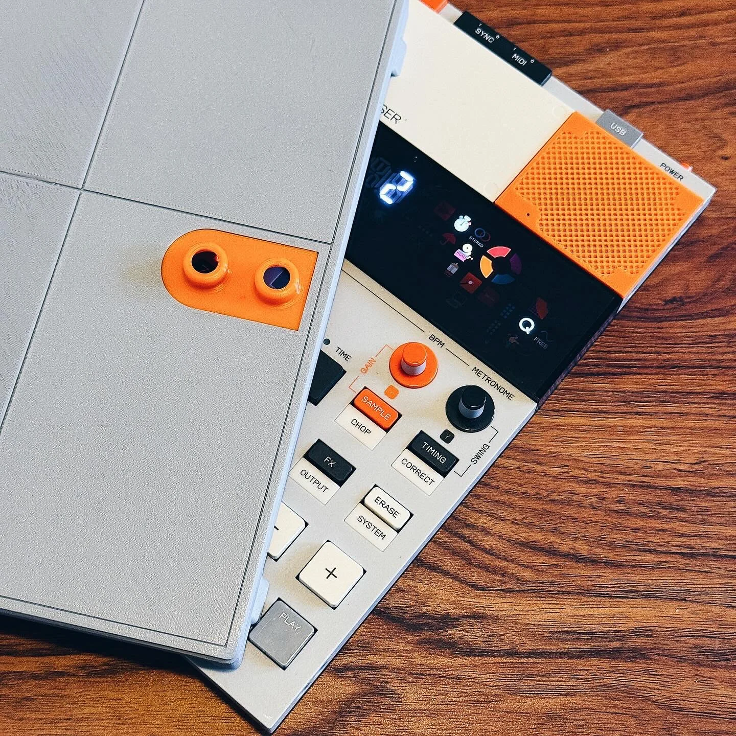 Close-up of an electronic drum machine and part of a laptop computer on a wooden surface. The drum machine has various controls, knobs, and a display screen, with orange accents.