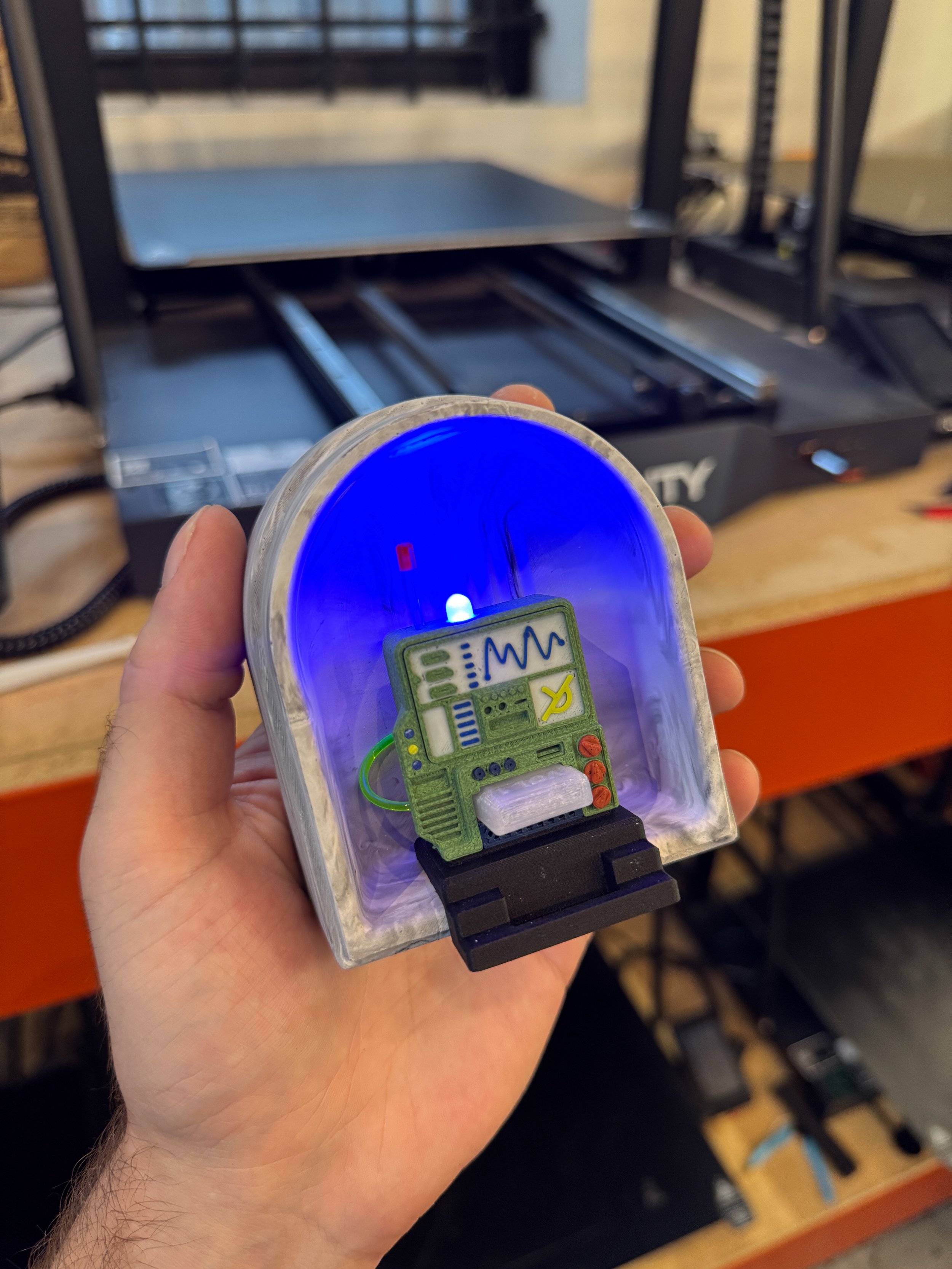 A person holding a small toy or model of a green robot with a digital display on its chest, in front of a 3D printer in a workshop or maker space.