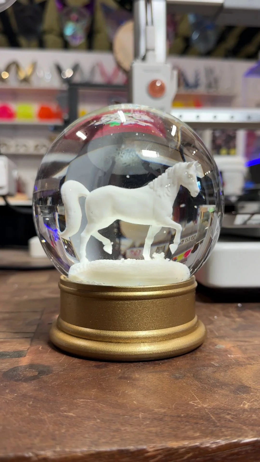 A snow globe featuring a white horse inside, placed on a wooden surface with a blurred store background.