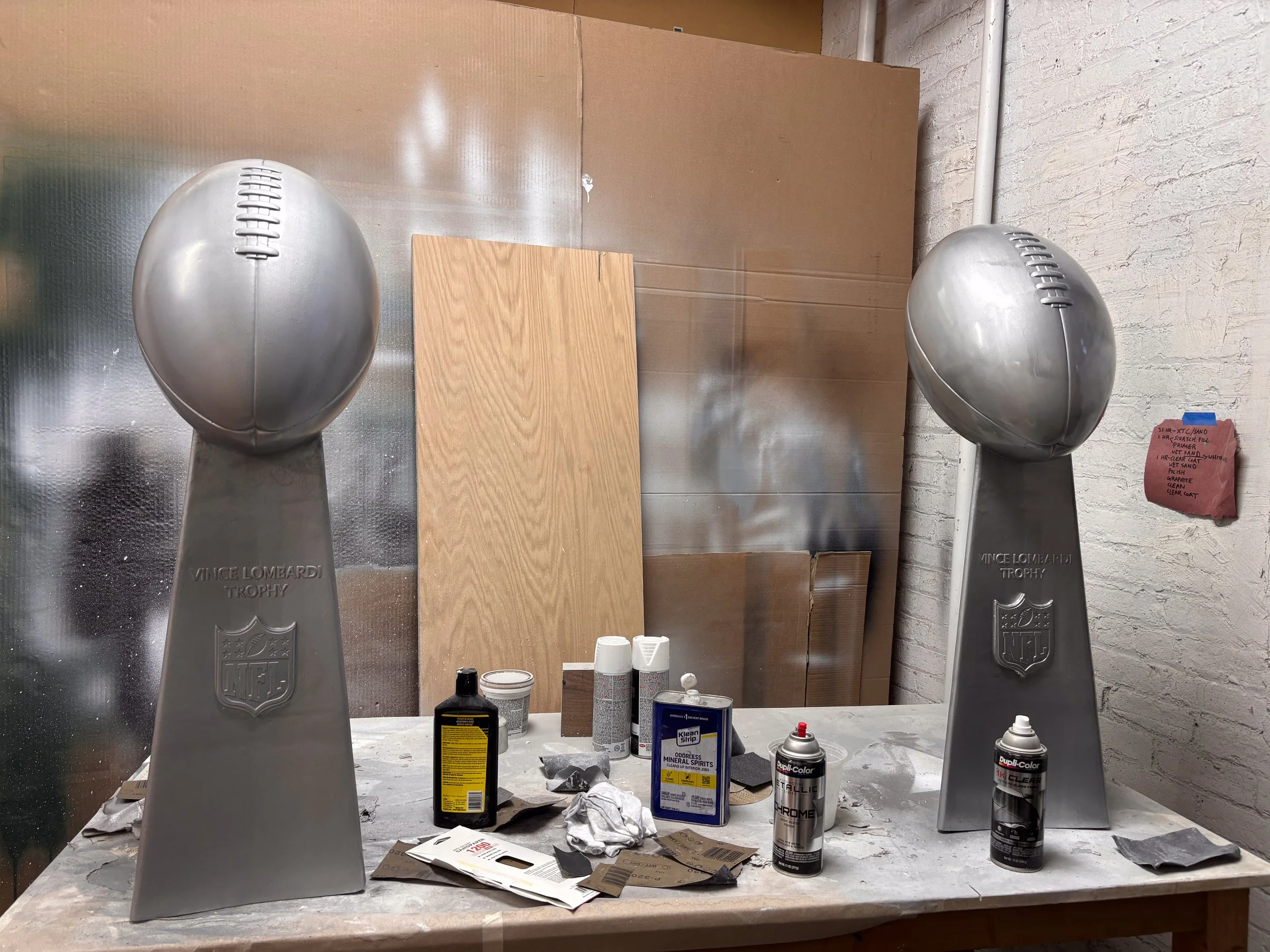 Garage workspace with two silver football-shaped NFL Vince Lombardi Trophy replicas, various spray paint cans, bottles, and supplies on a cluttered table, with a cardboard backdrop and a white brick wall.