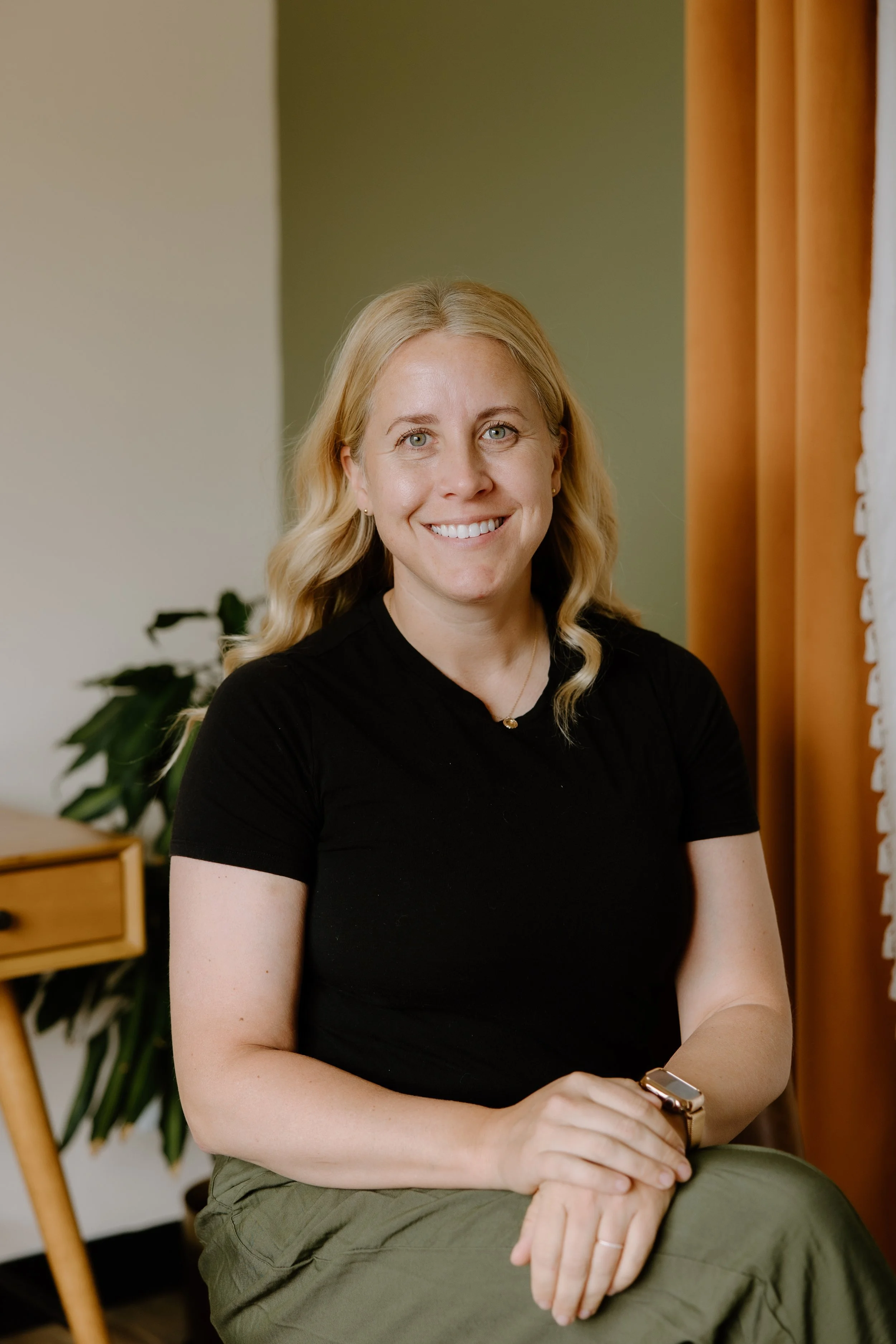 A woman with blonde hair and blue eyes smiling while standing in front of a wooden wall art piece in a beige room.