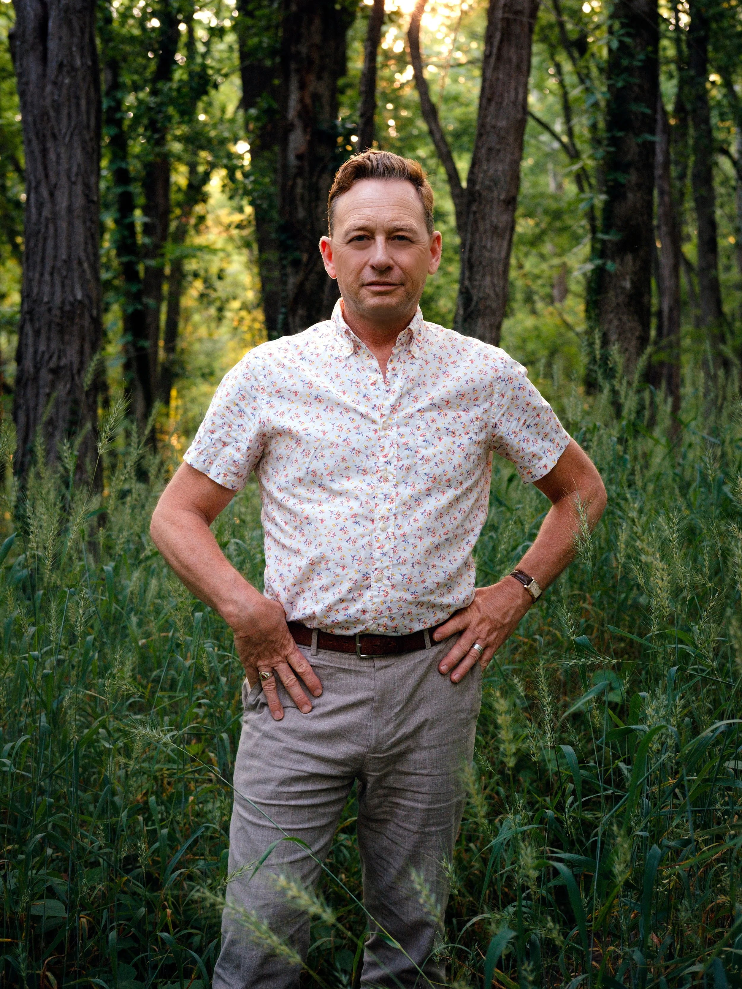 Silas House, a white man wearing a flowered white shortsleeve button up shirt with his hands on his hips in a forest.