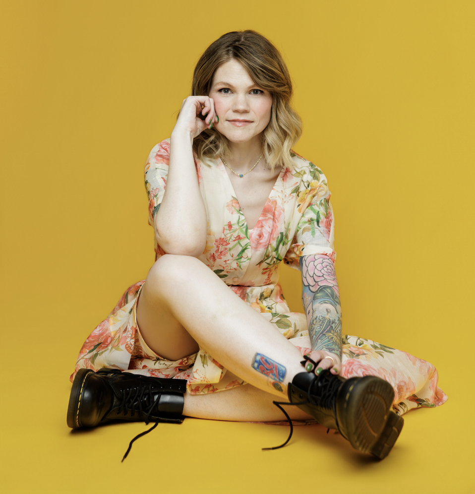 Photo of Mollie Hawkins in a floral dress and combat boots against a yellow-gold background.