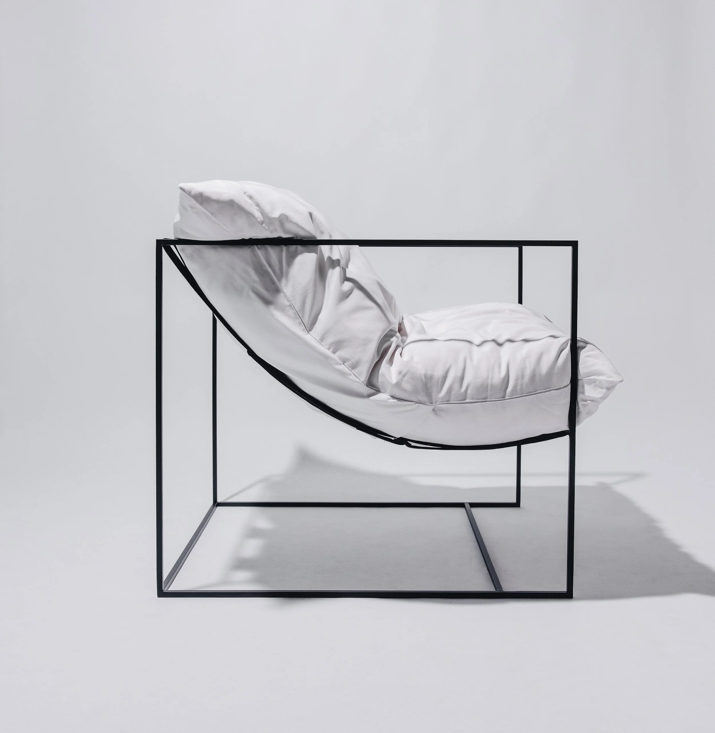 A chair cushion inside a black metal frame with white pillows and bedding, set against a plain white background.