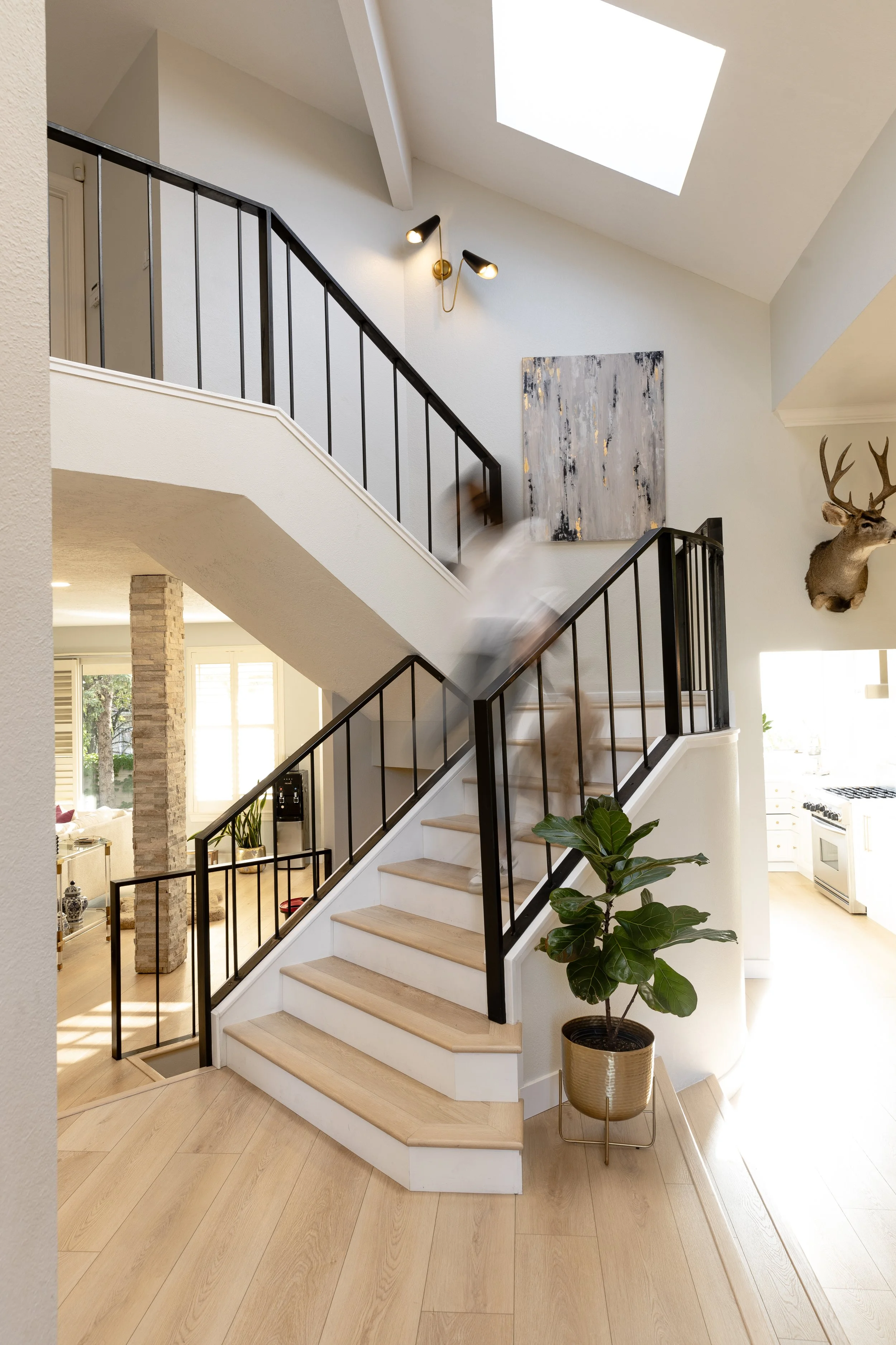 Photo of a modern interior staircase with light wooden steps, black metal railing, and a potted plant at the base. A person is blurred mid-motion going up, with a mounted deer head, artwork, and wall-mounted lights visible on the wall. A skylight brings natural light into the space.