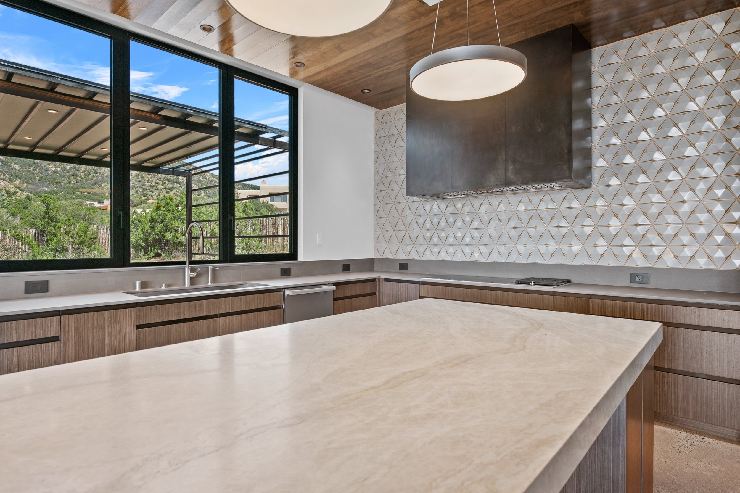 Modern kitchen with large window, wooden cabinets, gray countertops, a geometric tiled backsplash, a stainless steel range hood, and a circular hanging light fixture.