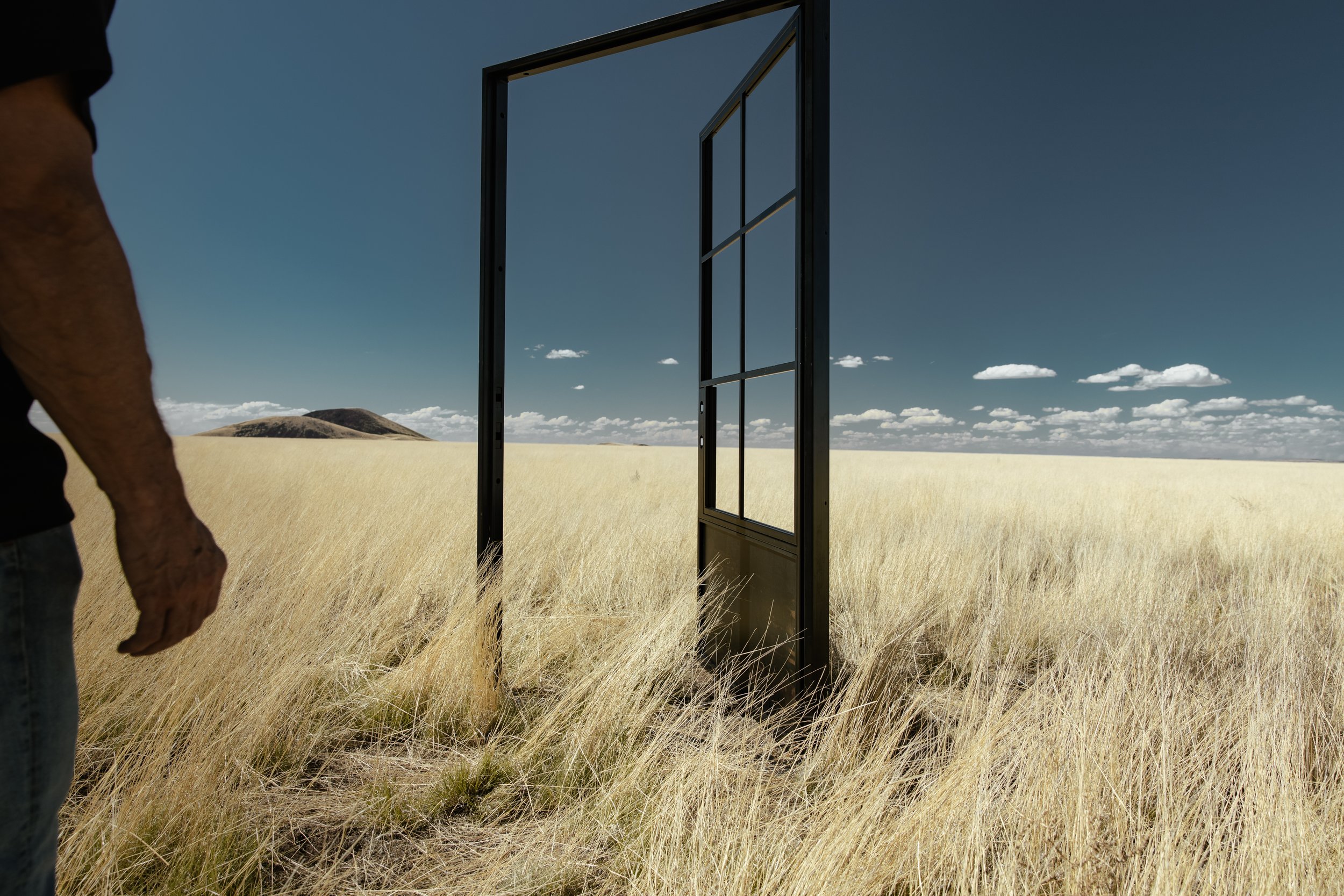 A person standing in a field of tall grass next to a large black door frame in the middle of the grass.
