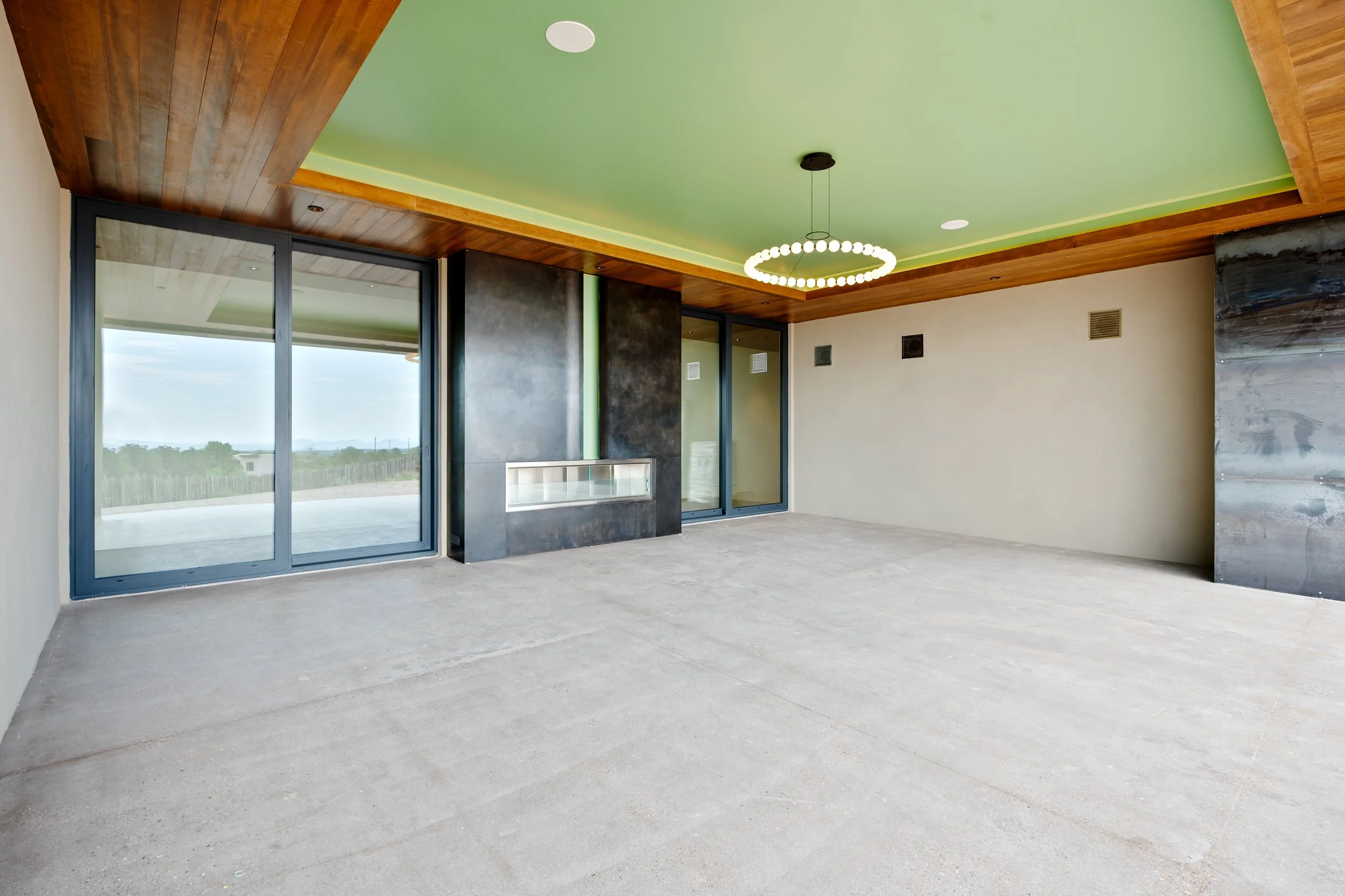 Empty room with large sliding glass doors, green ceiling with wooden border, modern black fireplace, circular chandelier, and concrete floor.