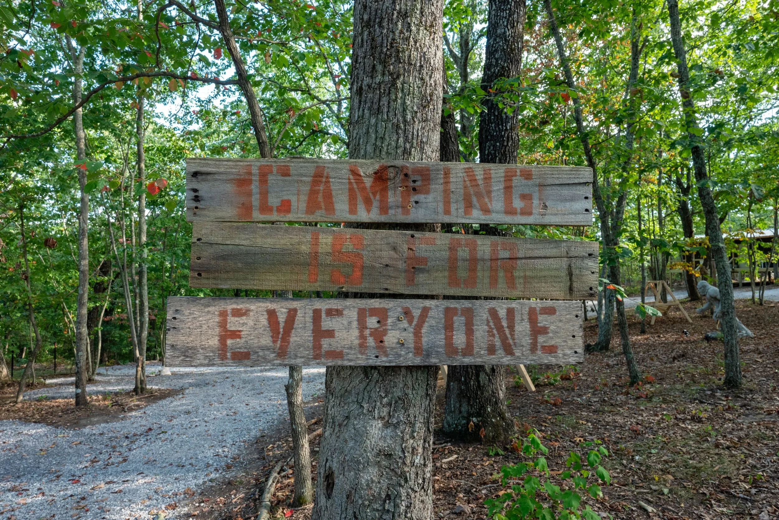 A wooden sign affixed to a tree reads 'Camping is for everyone' in red painted letters. The sign is in a wooded outdoor setting with trees and a gravel path visible.