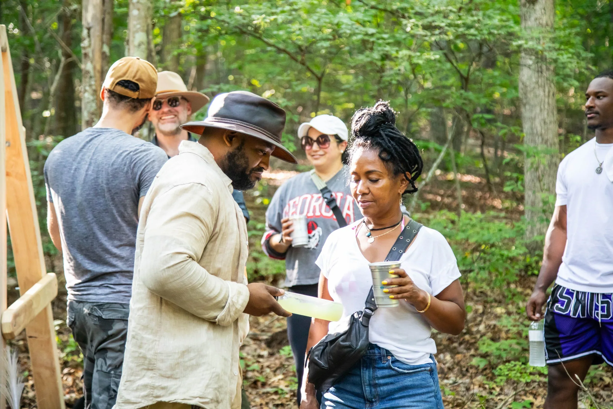 Group of six diverse people standing and talking in a wooded outdoor area, some holding cups or water bottles, engaging in a social gathering.