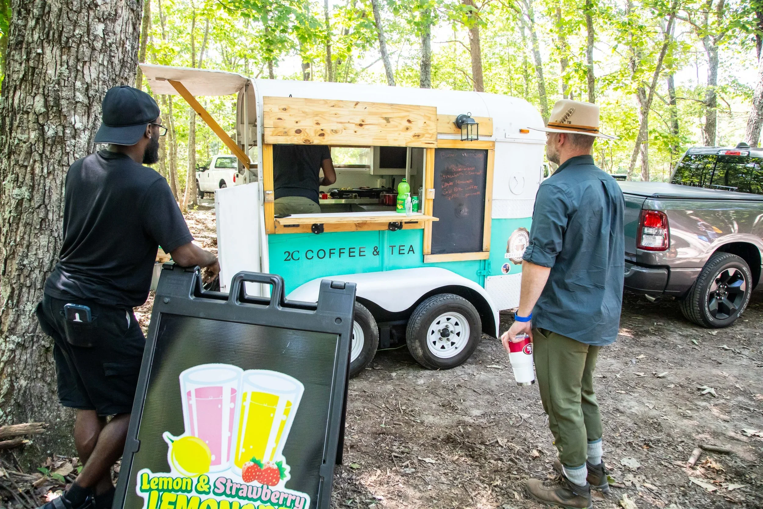 A food truck labeled "2C Coffee & Tea" parked in a wooded area, serving two customers. One man, wearing a black cap and black shirt, stands at the window, while another man, wearing a wide-brimmed hat, dark green pants, and a blue shirt, holds a cup.