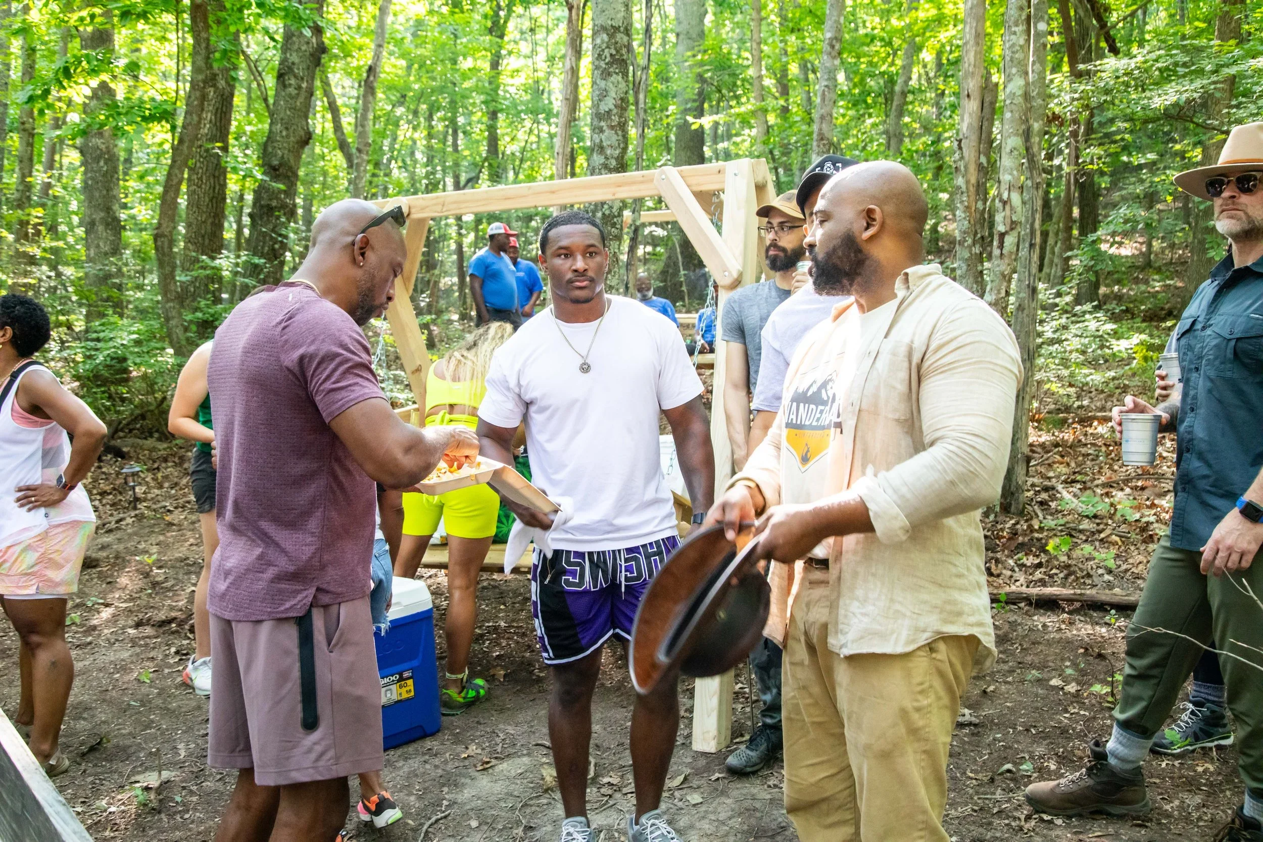 Group of people gathered in a forest for an outdoor event or picnic, with some holding food and drinks, and others conversing.