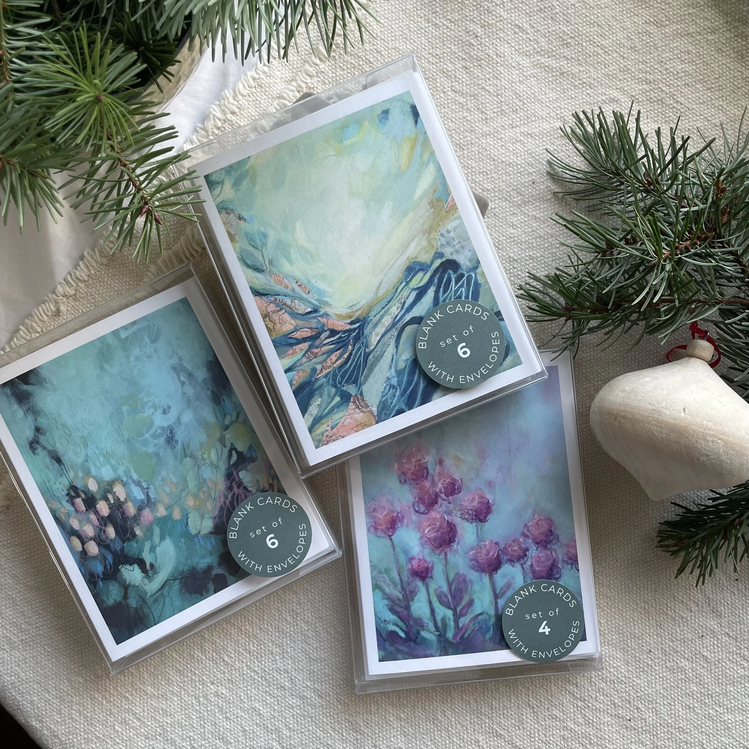 Three packs of blank greeting cards with abstract paintings on the covers, each pack containing 4 or 6 cards, placed on a table with pine branches and a wooden ornament nearby.