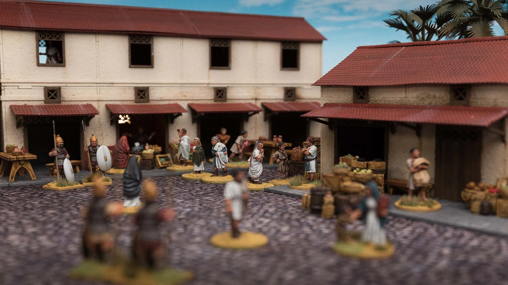 Miniature diorama of a bustling ancient market featuring painted Sarissa Precision MDF model buildings, civilian figures, and merchants.