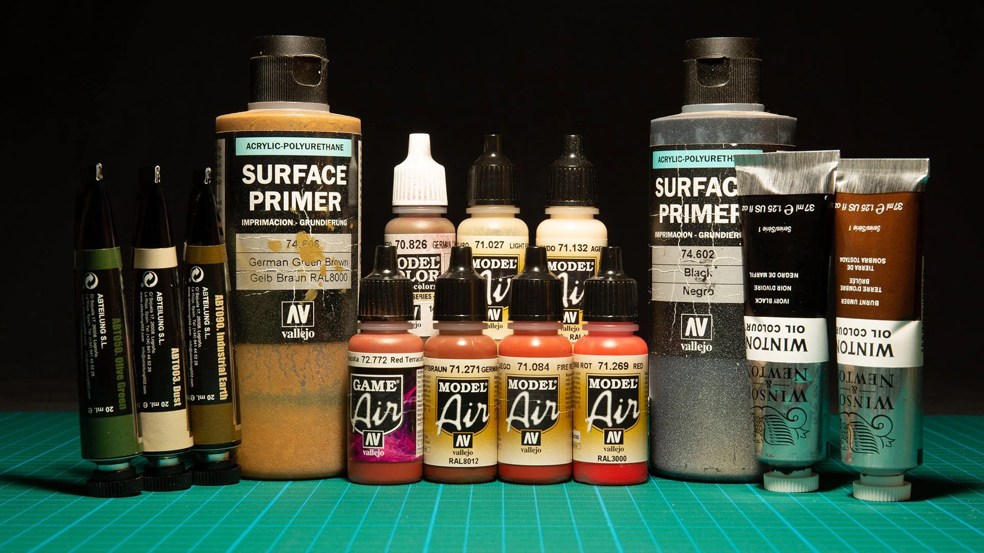 Various miniature modeling supplies on a green mat, including Vallejo surface primers, acrylic paints, and oil colors for weathering.