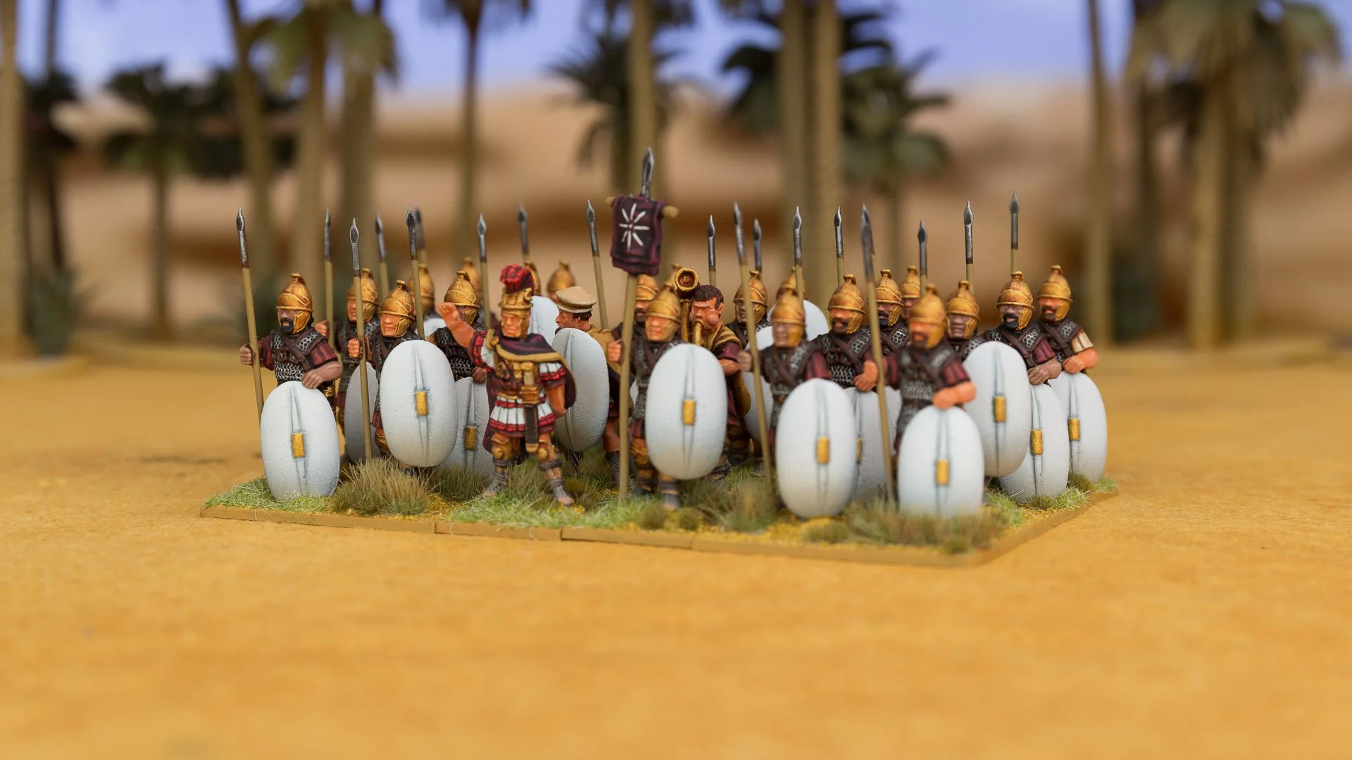 Regiment of painted miniature Ptolemaic infantry holding spears and large white oval shields in a desert setting.