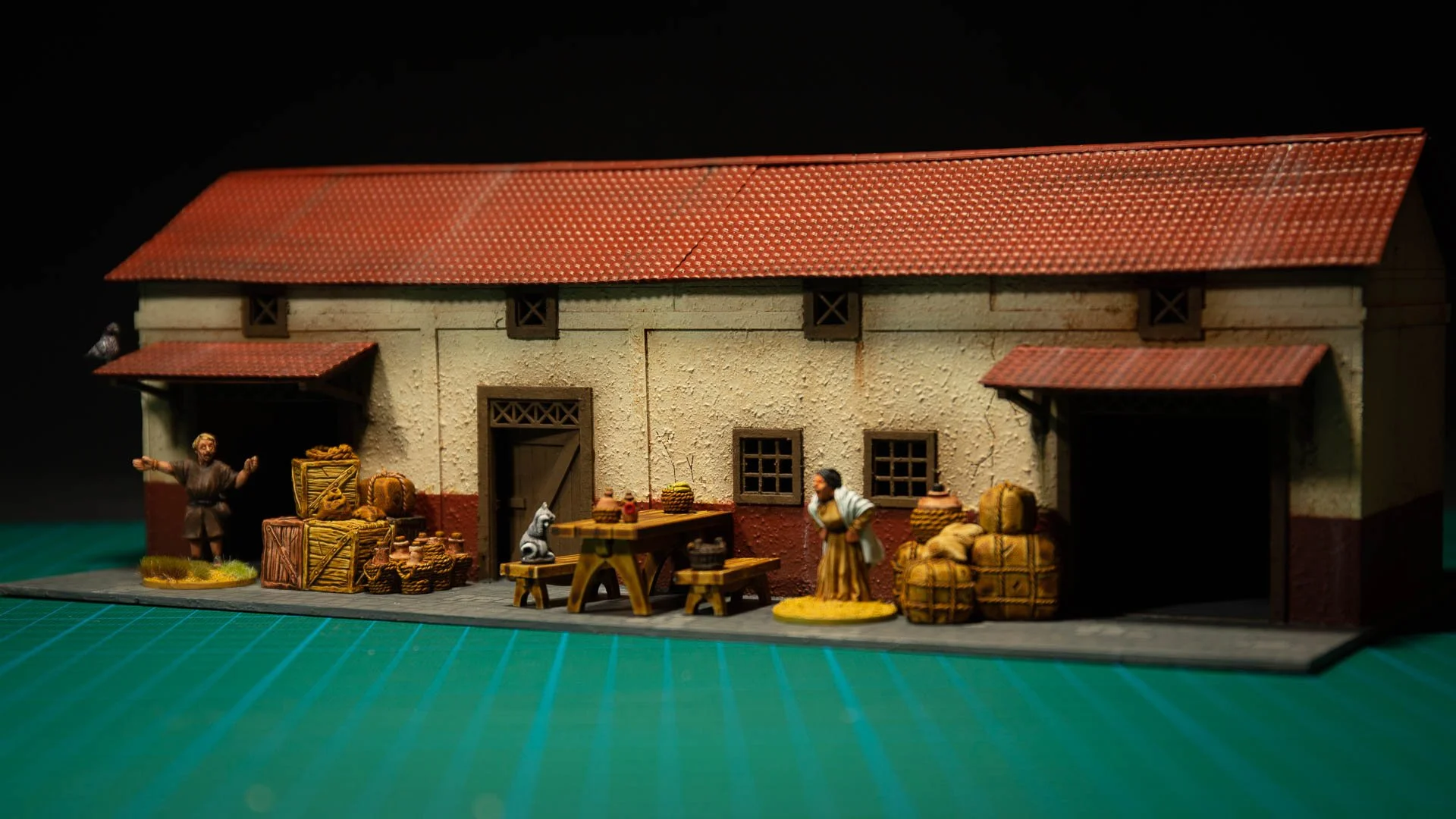 Painted MDF model warehouse building decorated with miniature diorama scatter terrain, including wooden crates, amphorae, tables, and civilian figures.