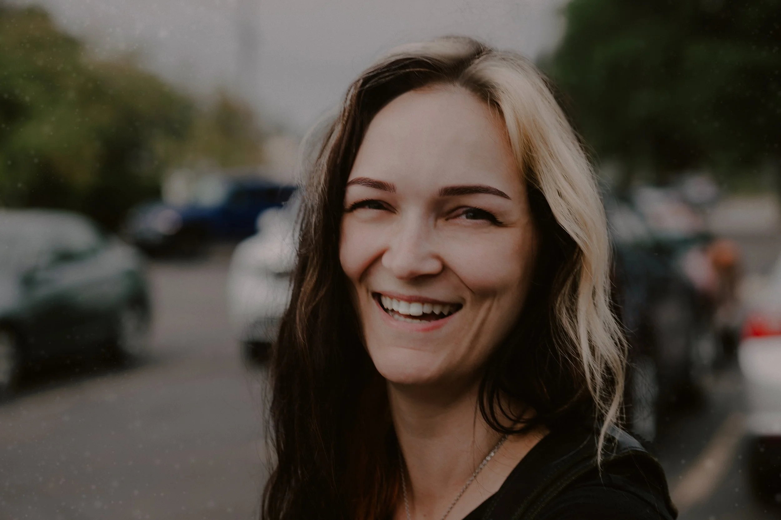 A woman with long dark hair and blonde highlights smiling and winking outdoors in a parking lot on a rainy day.