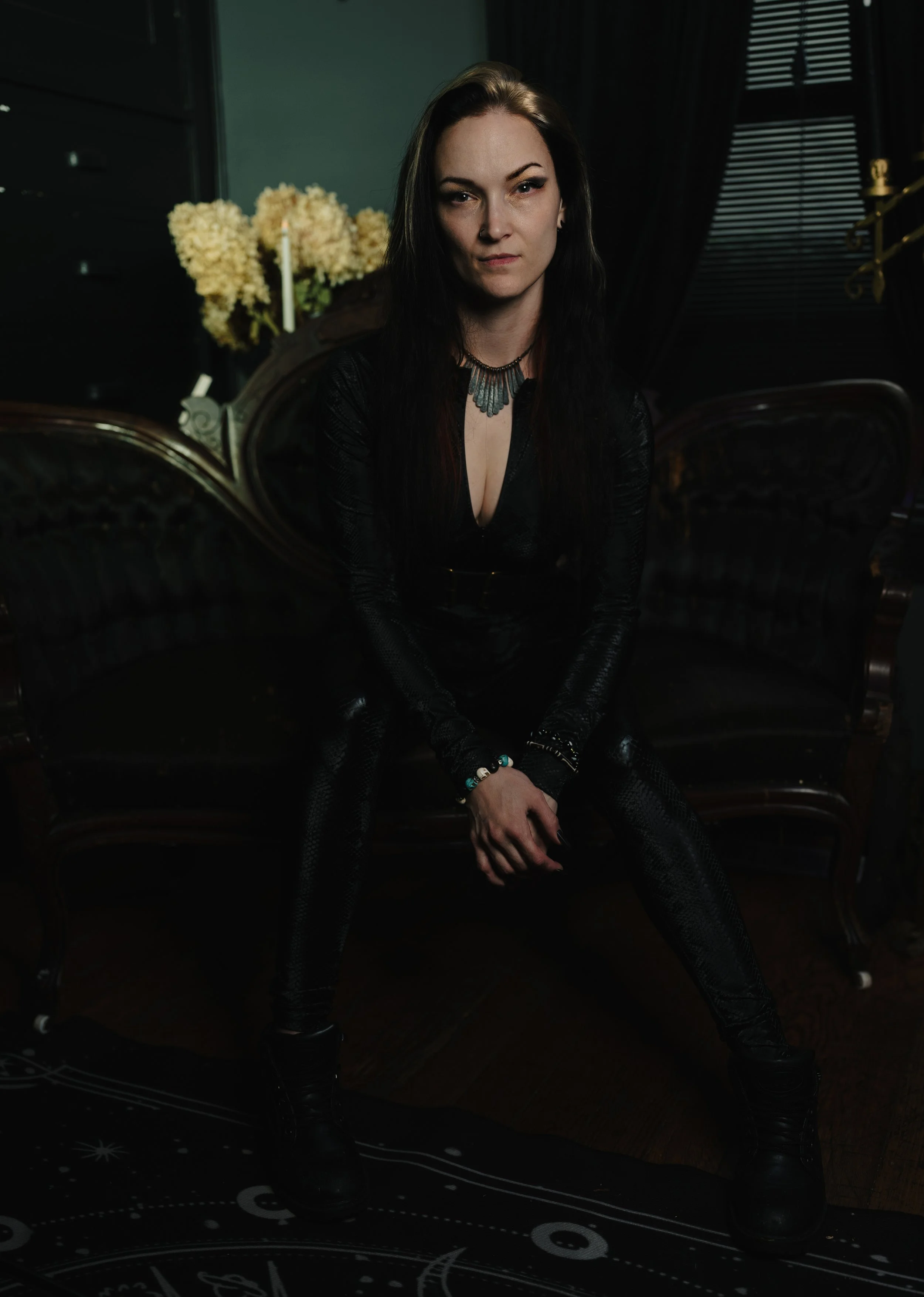 A woman with dark hair and light skin wearing a black top with a deep neckline and jewelry, sitting on a black vintage-style sofa in a dimly lit room with dark curtains and a window with closed blinds behind her. A bouquet of cream-colored flowers an