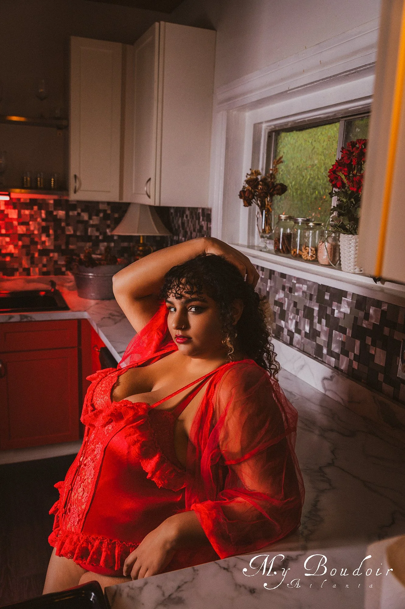 Person in red lingerie and robe posing in a kitchen setting with a marble countertop and mosaic backsplash.