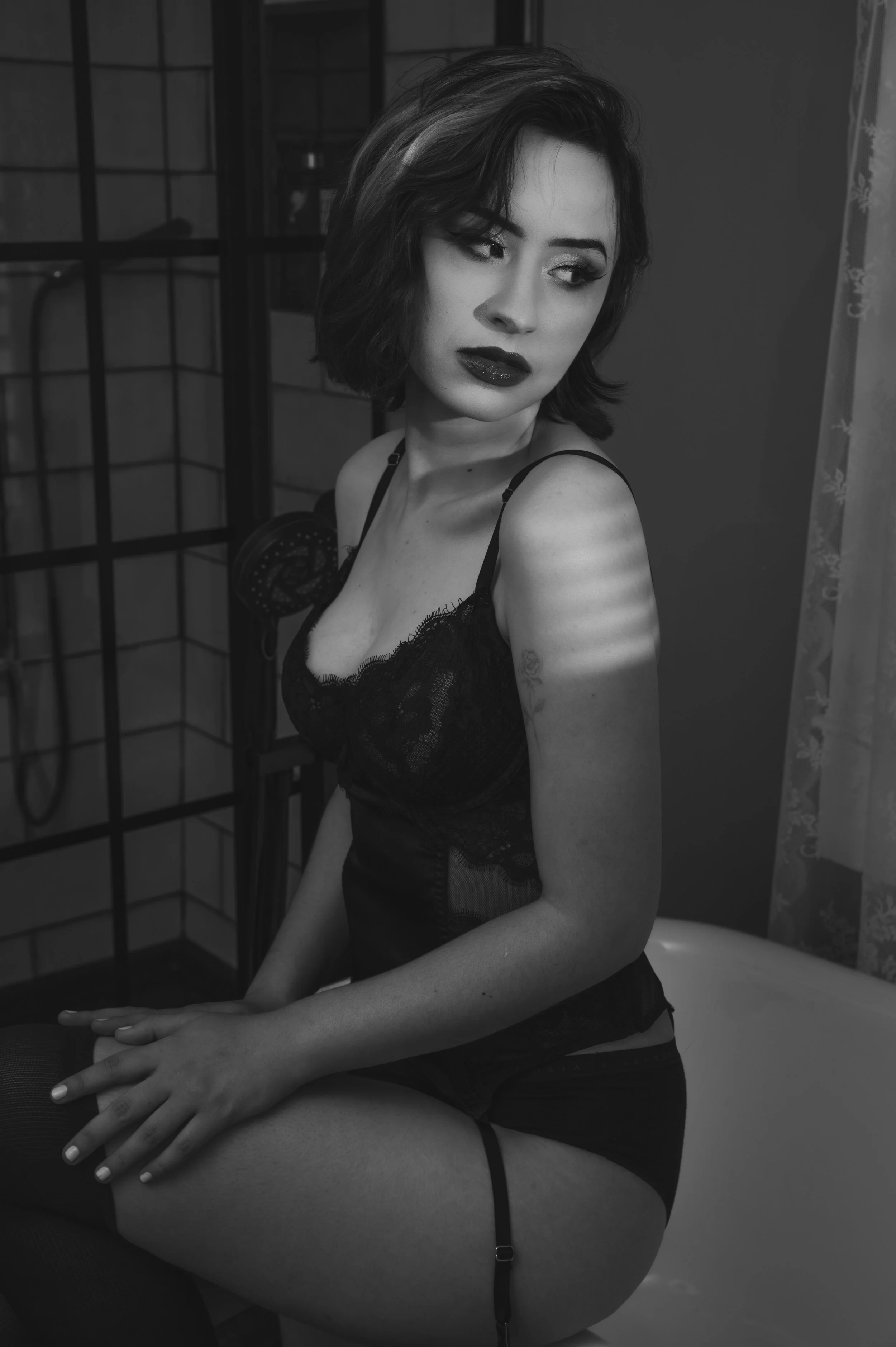 Black and white photo of a woman in a lace outfit sitting on a bathtub, looking thoughtfully to the side.