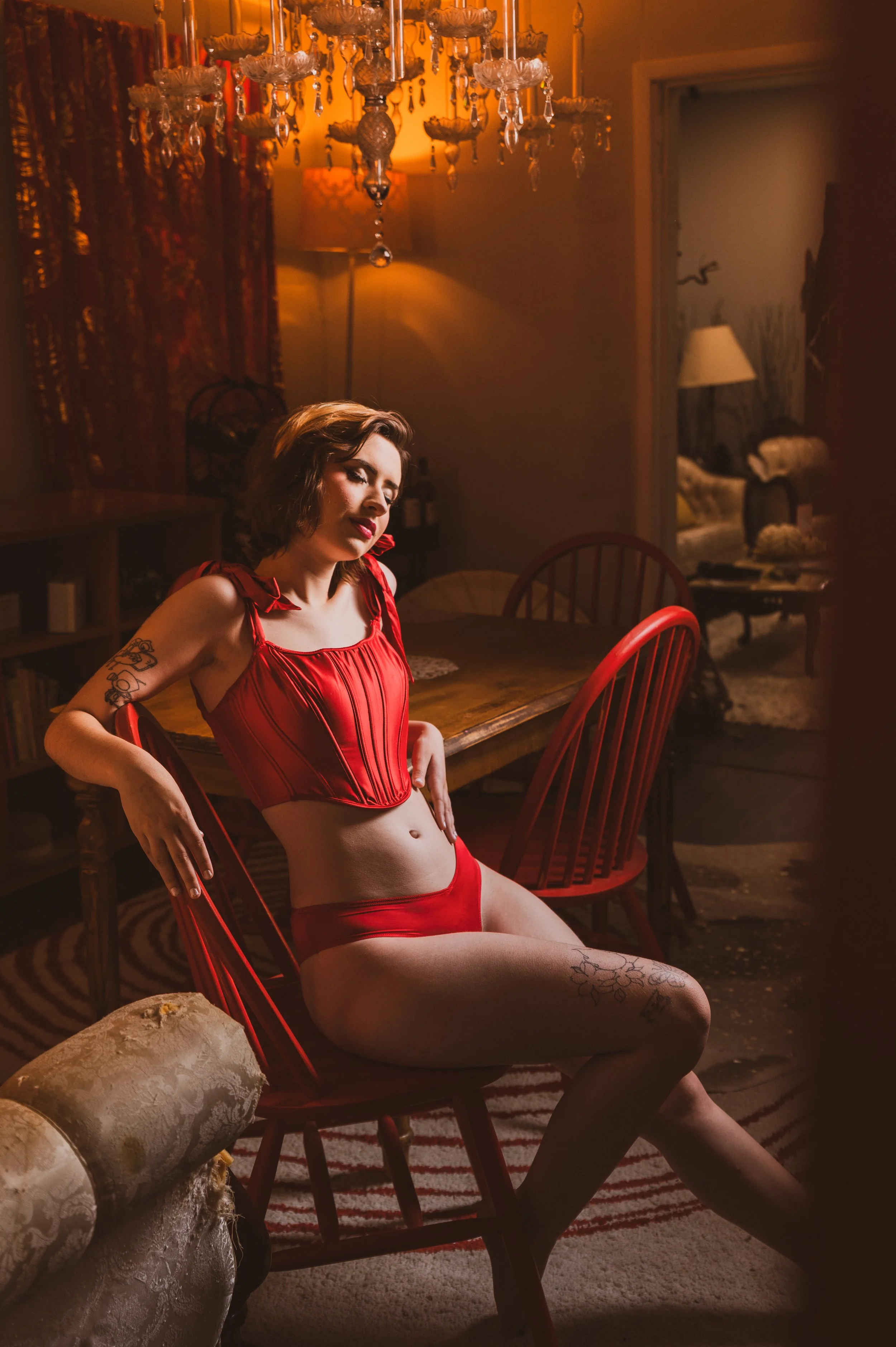 Person in red lingerie sitting on a chair in a dimly-lit room with chandelier and vintage decor.