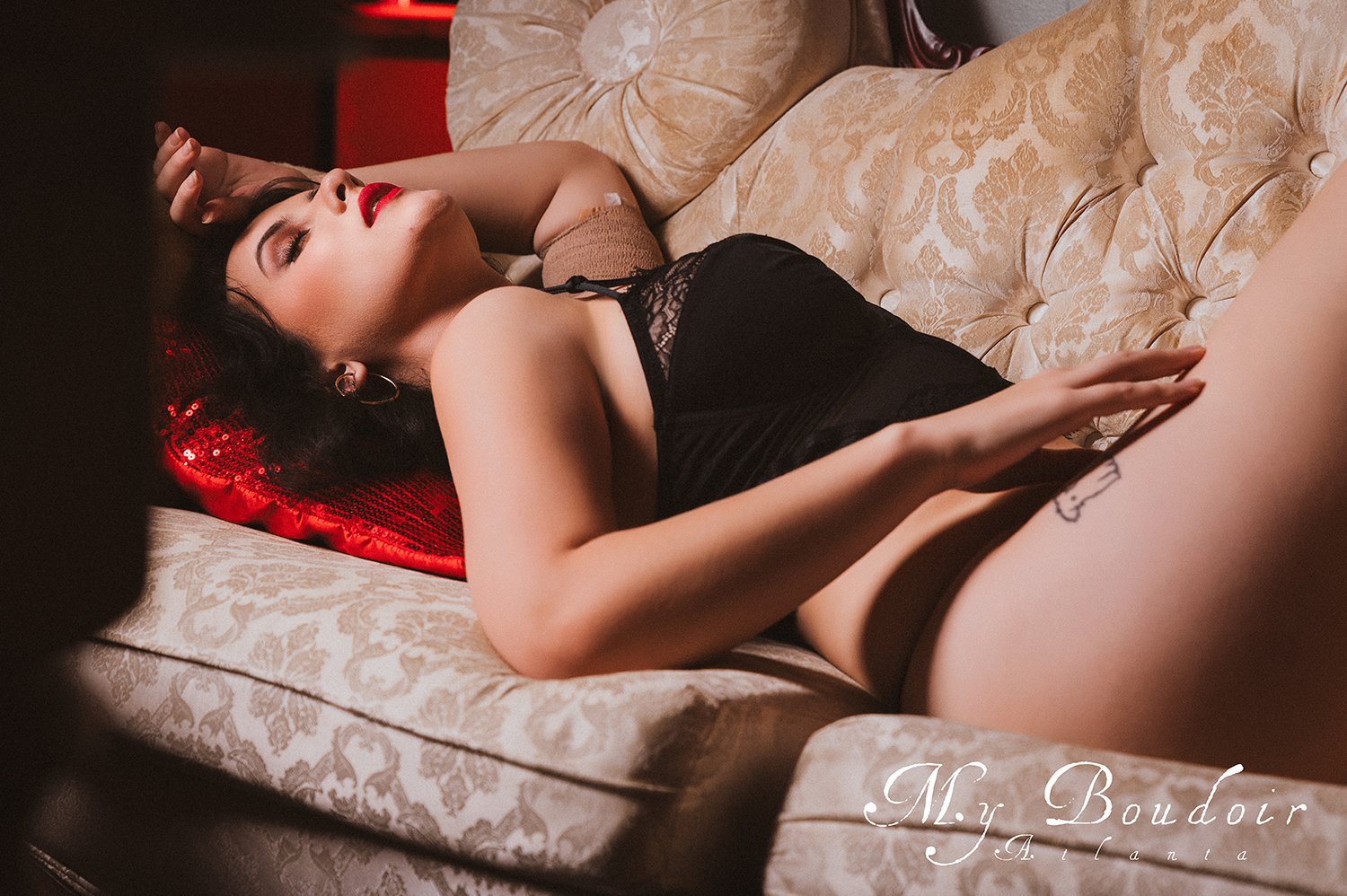 Person in black lingerie reclining on a plush, patterned sofa with red sequin pillow.