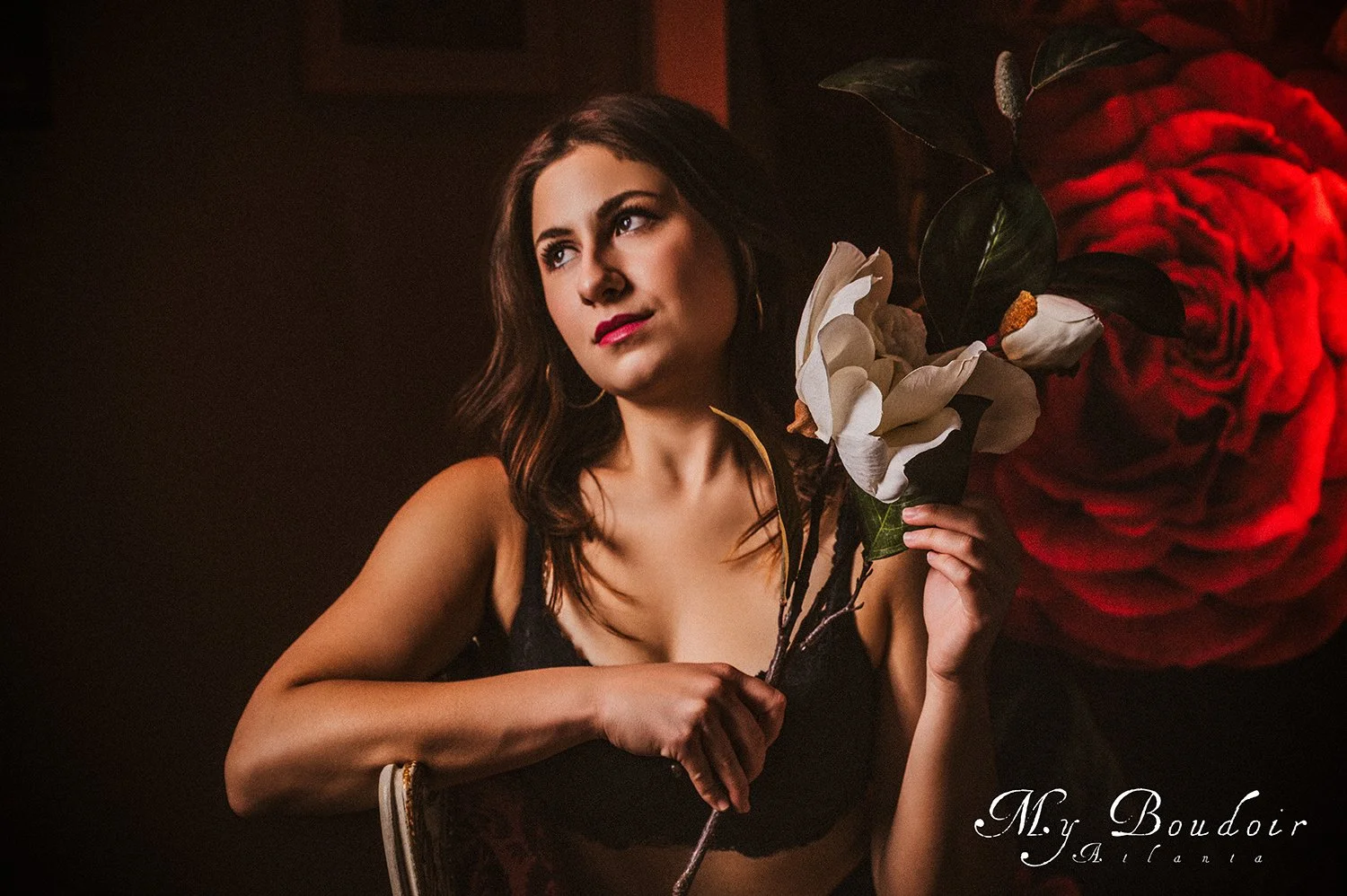 Woman in black lingerie holding a large white flower, sitting in a dimly lit room with a red rose background, "My Boudoir Atlanta" label in the corner.