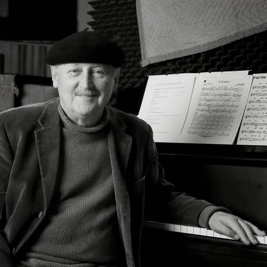 An older man wearing a beret and sweater sitting at a piano with sheet music, in a recording studio with soundproofing walls.