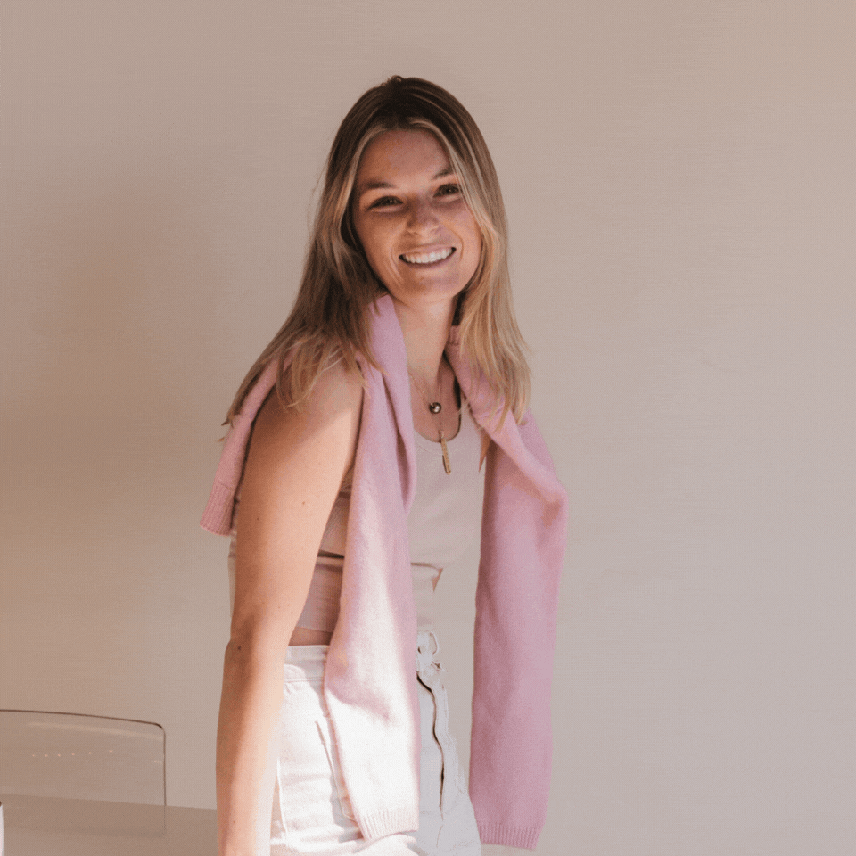 Delaney from Carefully Counted with blonde hair smiling, wearing a white top, beige pants, and a pink sweater draped over her shoulders, standing against a plain background.