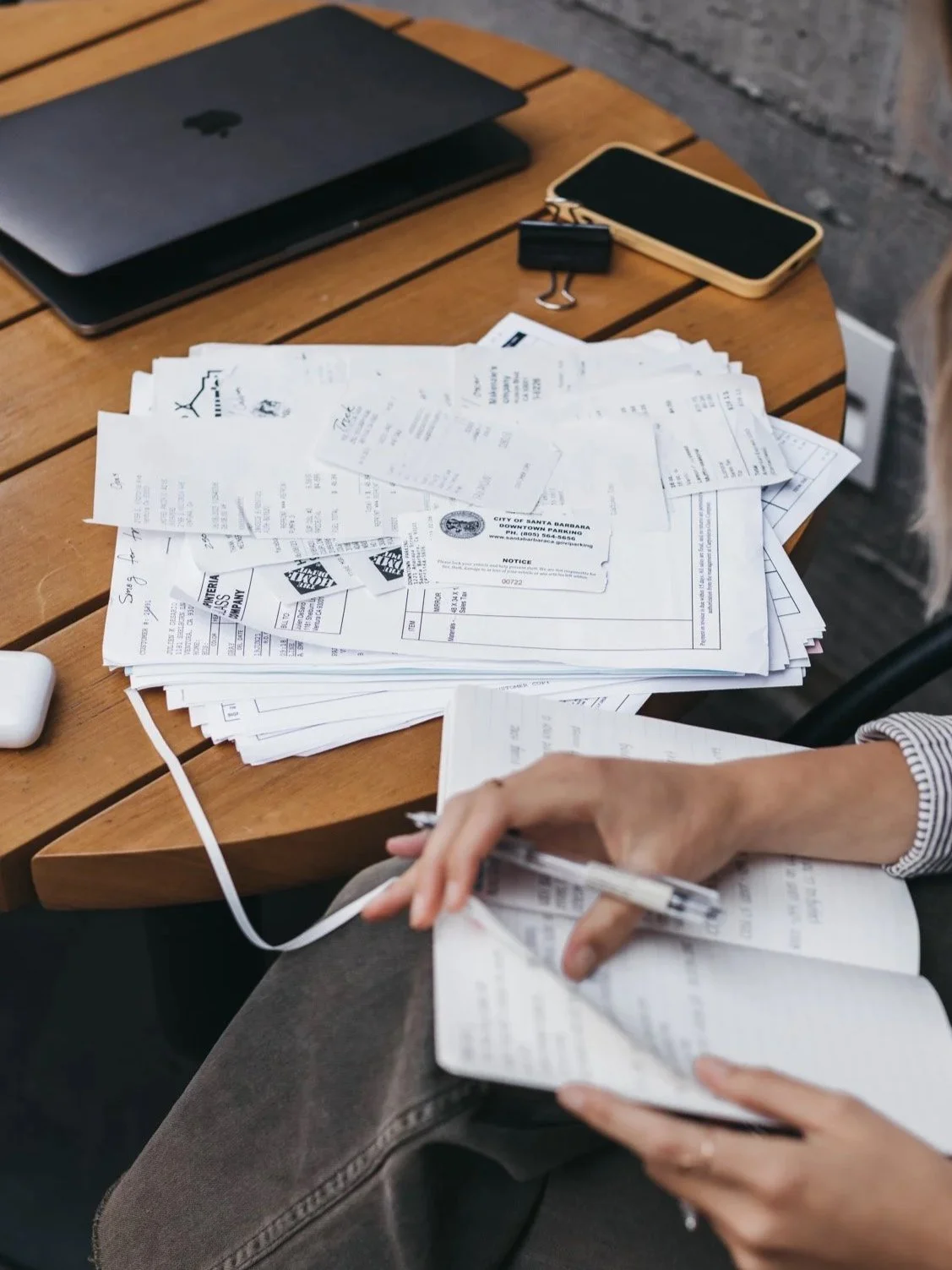 Person organizing paper receipts at a wooden table with a laptop, smartphone, and binder clip.