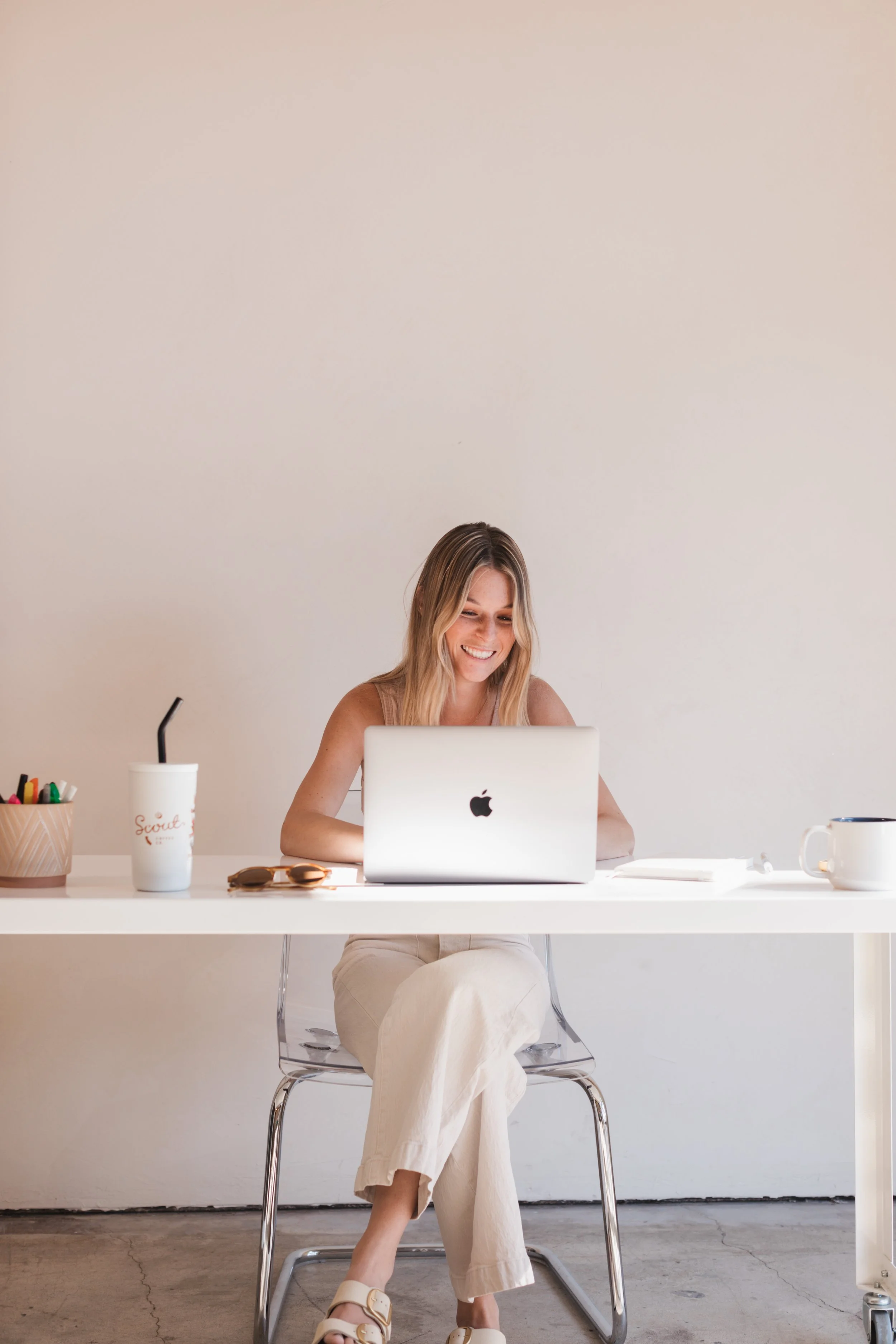 A young woman smiling while working on a laptop at a white desk, with a disposable cup, sunglasses, pen holder, and mug on the desk prepping taxes in San Luis Obispo.