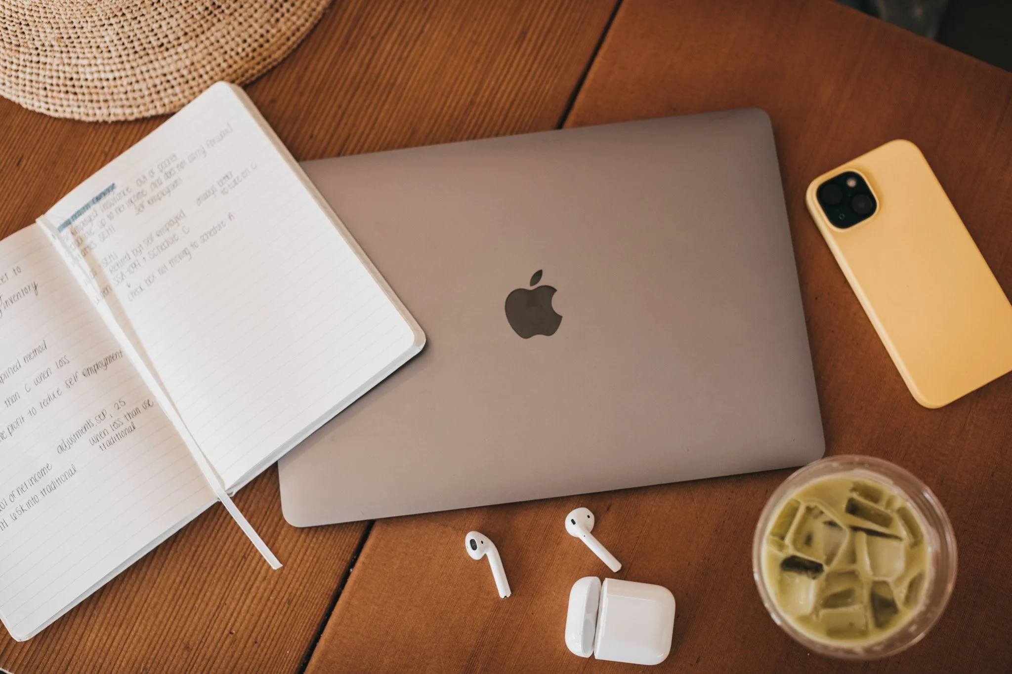 A laptop, open notebook, iPhone, wireless earbuds, and iced coffee on a wooden table.